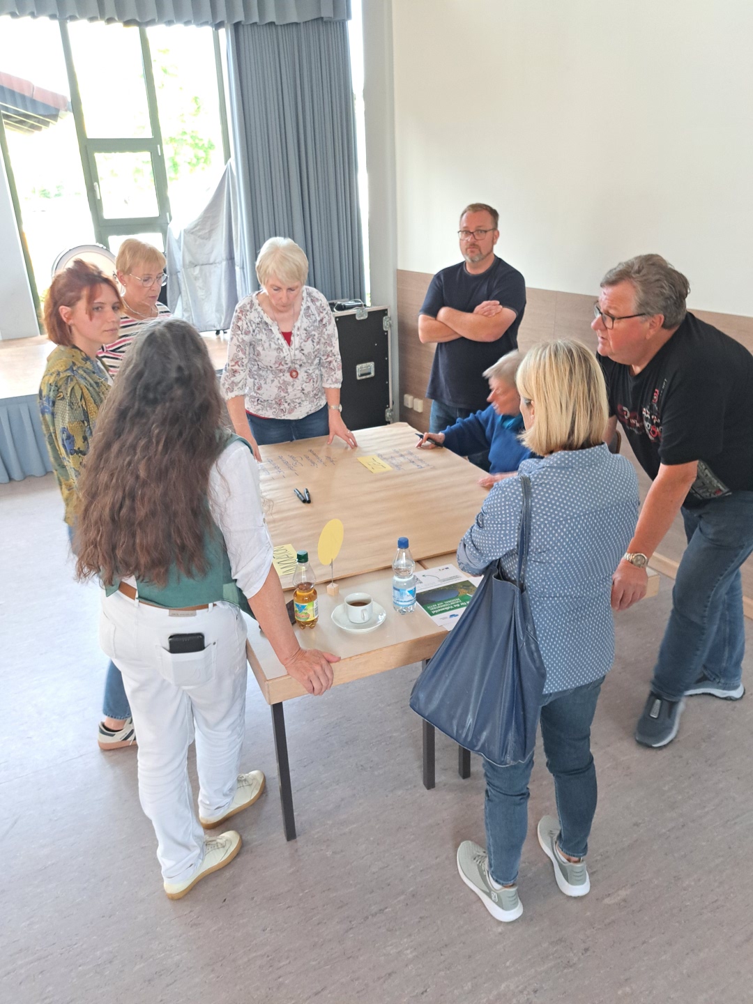 Sieben Männer und Frauen stehen um einen Tisch und tauschen sich aus. Auf dem Tisch liegen Zettel und Stifte sowie eine Tasse und zwei Getränke. Die Menschen tauschen sich zum Projekt VereinsKompass aus.