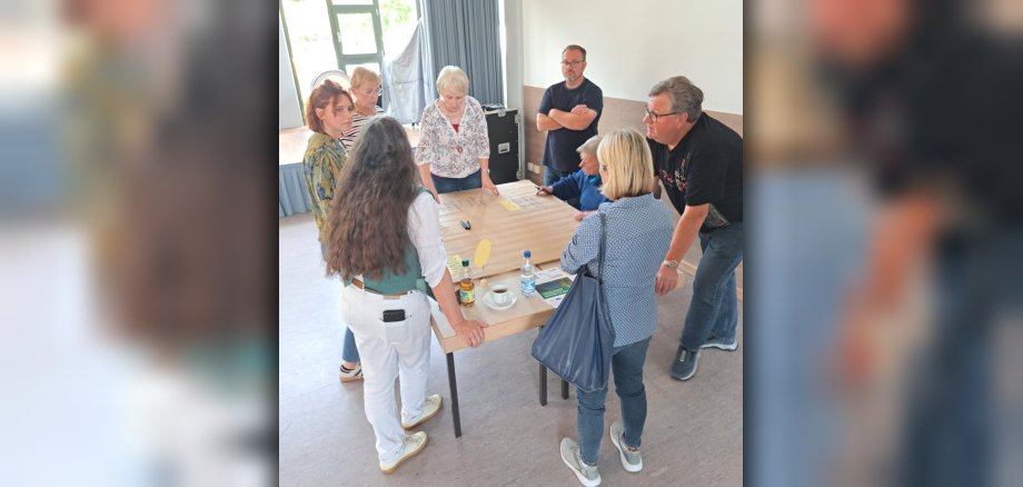 Sieben Männer und Frauen stehen um einen Tisch und tauschen sich aus. Auf dem Tisch liegen Zettel und Stifte sowie eine Tasse und zwei Getränke. Die Menschen tauschen sich zum Projekt VereinsKompass aus.