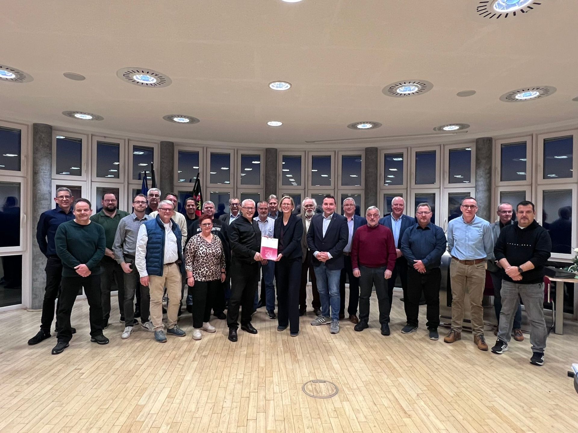 Vor einer Fensterfront stehen Bürgermeister Albert Jung und Staatsekretärin Simone Schneider (Mitte) und halten den Förderbescheid in den Händen. Drum herum stehen der Stadtbürgermeister und die Ortsbürgermeistern sowie Beigeordneten der Gemeinden. 