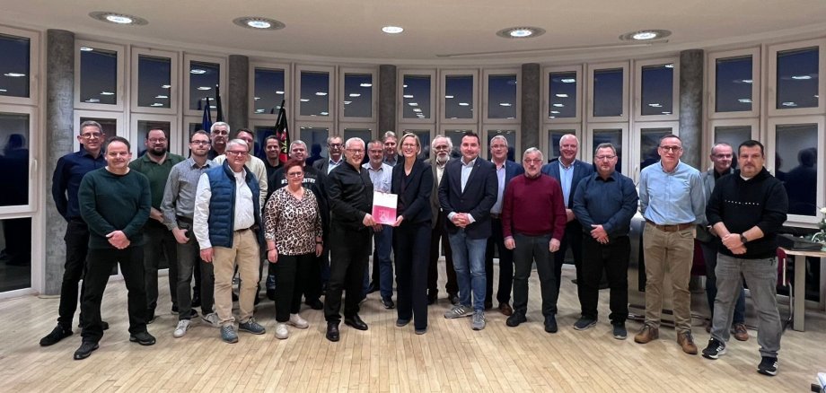 Bürgermeister Albert Jung und Staatsekretärin Simone Schneider (Mitte) freuen sich mit dem Stadtbürgermeister und den Ortsbürgermeistern sowie Beigeordneten der Gemeinden über die Förderung. Vor einer Fensterfront stehen Bürgermeister Albert Jung und Staatsekretärin Simone Schneider (Mitte) und halten den Förderbescheid in den Händen. Drum herum stehen der Stadtbürgermeister und die Ortsbürgermeistern sowie Beigeordneten der Gemeinden.