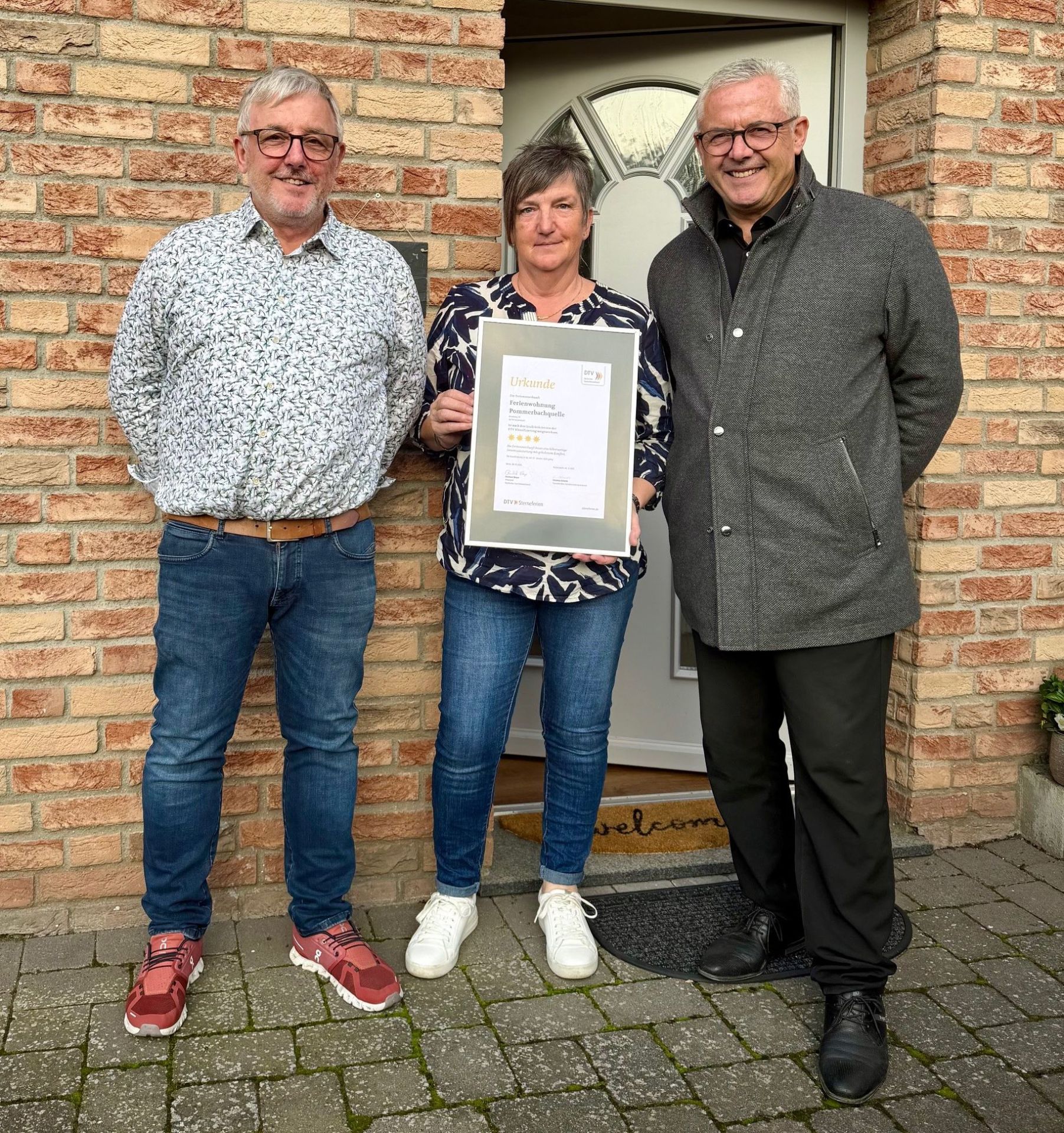 Vor dem Hauseingang der Ferienwohnung Pommerbachquelle stehen Herbert und Petra Peters mit Bürgermeister Albert Jung. Frau Peters hält einen Bilderrahmen mit der Klassifizierungsurkunde in den Händen. 
