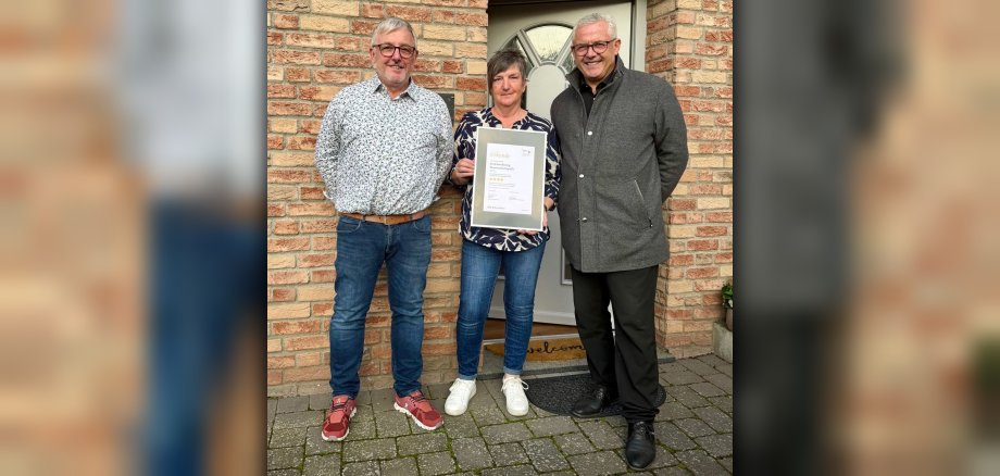 Vor dem Hauseingang der Ferienwohnung Pommerbachquelle stehen Herbert und Petra Peters mit Bürgermeister Albert Jung. Frau Peters hält einen Bilderrahmen mit der Klassifizierungsurkunde in den Händen. 