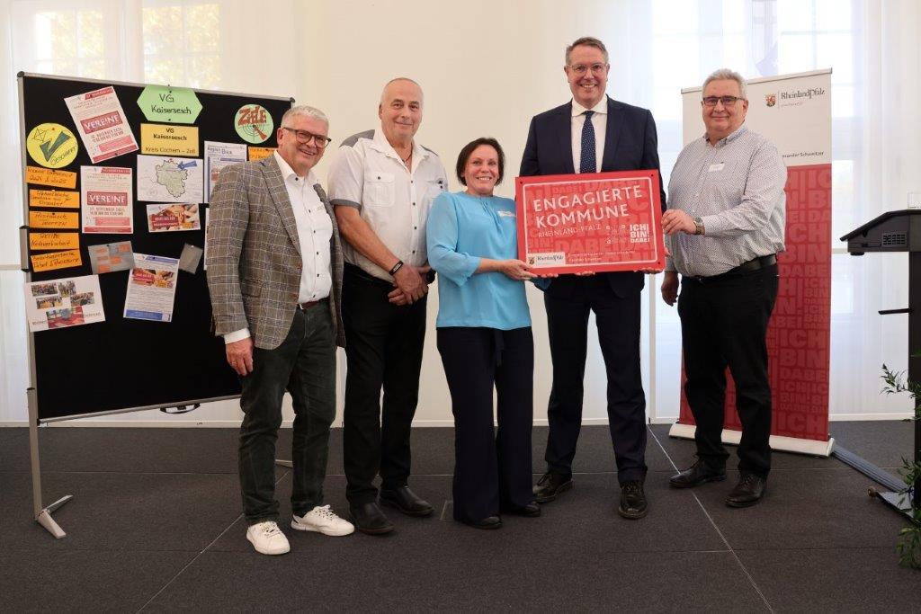 Vor einer Stellwand, die mit vielen unterschiedlichen Plakaten bestückt ist und einem Roll-up der Staatskanzlei Rheinland-Pfalz, stehen von links nach rechts: Erster Beigeordneter Gerhard Weber, Hans Günter Labonte, Bianca Sesterhenn, Ministerpräsident Alexander Schweitzer, Norbert Fuhrmann. Bianca Sesterhenn und Ministerpräsident Alexander Schweitzer halten gemeinsam ein rotes Schild mit der Aufschrift "Engagierte Kommune" in ihren Händen.