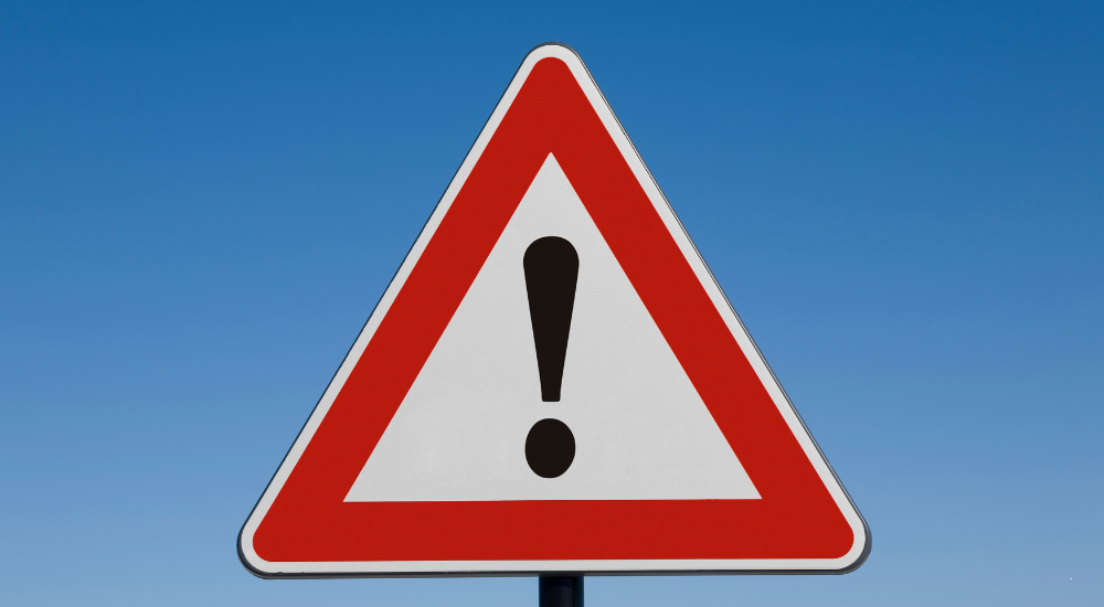 Vor einem blauen Hintergrund, der den Himmel darstellen soll, ist ein dreieckiges Schild mit weißem und rotem Rand zu sehen. Das Schild ist ein Achtungsschild, in dem ein schwarzes Ausrufezeichen enthalten ist.