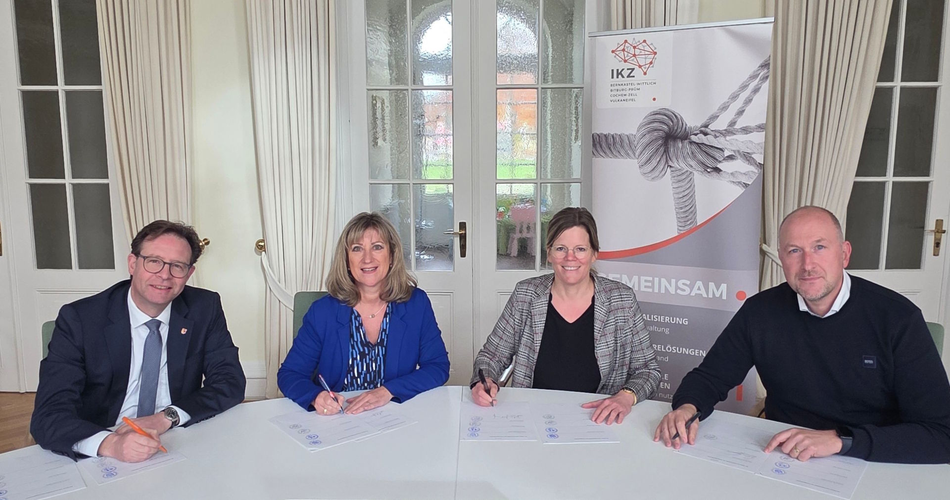 Landrätinnen und Landräte unterzeichnen die Kooperationsvereinbarung zum IKZ-Portal. An einem runden, weißen Tisch sitzen zwei Landrätinnen und zwei Landräte. Im Hintergrund sind Fenster und Vorhänge zu sehen. Am rechten Rand steht ein Roll-up mit dem IKZ-Portal-Logo. Darunter abgebildet sieht man ein Seil mit Knoten, das an den Enden in einzelnen Strängen auseinandergeht.