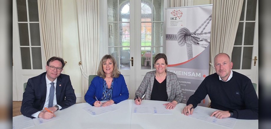Landrätinnen und Landräte unterzeichnen die Kooperationsvereinbarung zum IKZ-Portal. Landrätinnen und Landräte unterzeichnen die Kooperationsvereinbarung zum IKZ-Portal. An einem runden, weißen Tisch sitzen zwei Landrätinnen und zwei Landräte. Im Hintergrund sind Fenster und Vorhänge zu sehen. Am rechten Rand steht ein Roll-up mit dem IKZ-Portal-Logo. Darunter abgebildet sieht man ein Seil mit Knoten, das an den Enden in einzelnen Strängen auseinandergeht.