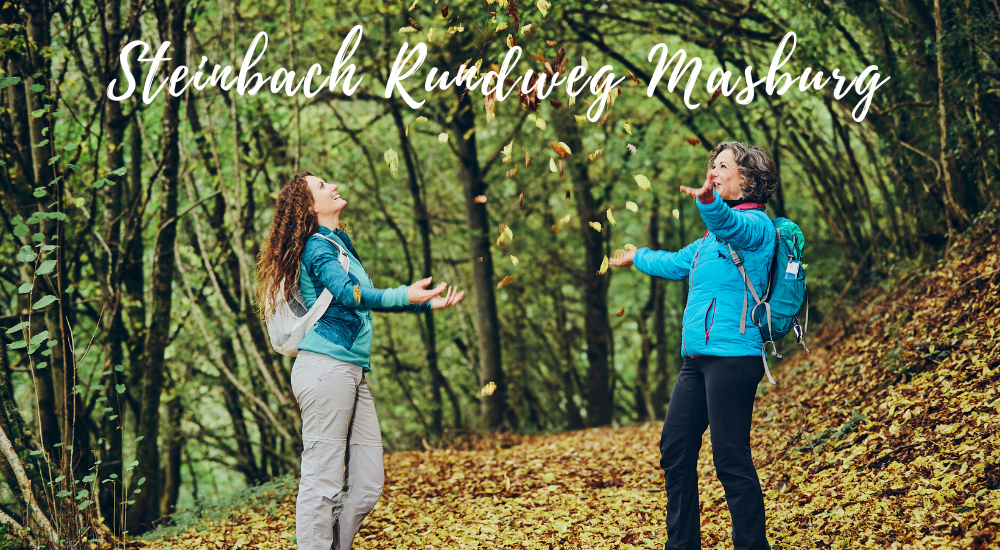 Zwei Frauen, die in einem herbstlichen Wald stehen, mit einem schriftlichen Hinweis auf den Steinbach-Rundweg Masburg.