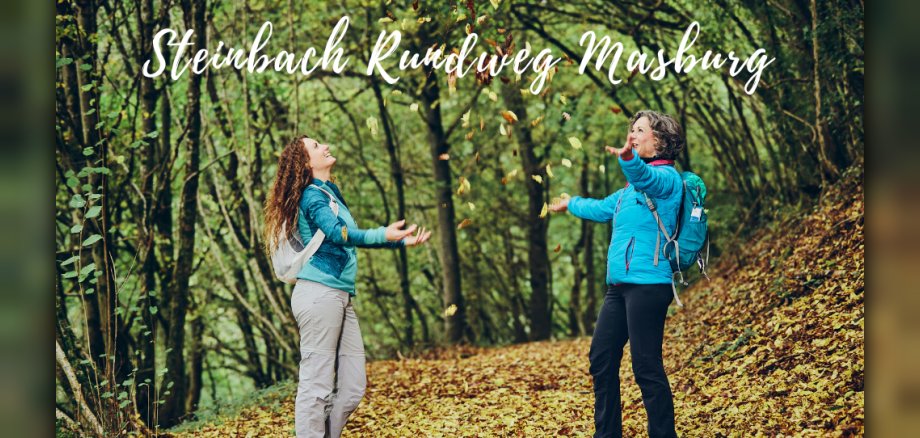 Zwei Frauen, die in einem herbstlichen Wald stehen, mit einem schriftlichen Hinweis auf den Steinbach-Rundweg Masburg. Zwei Frauen, die in einem herbstlichen Wald stehen, mit einem schriftlichen Hinweis auf den Steinbach-Rundweg Masburg.