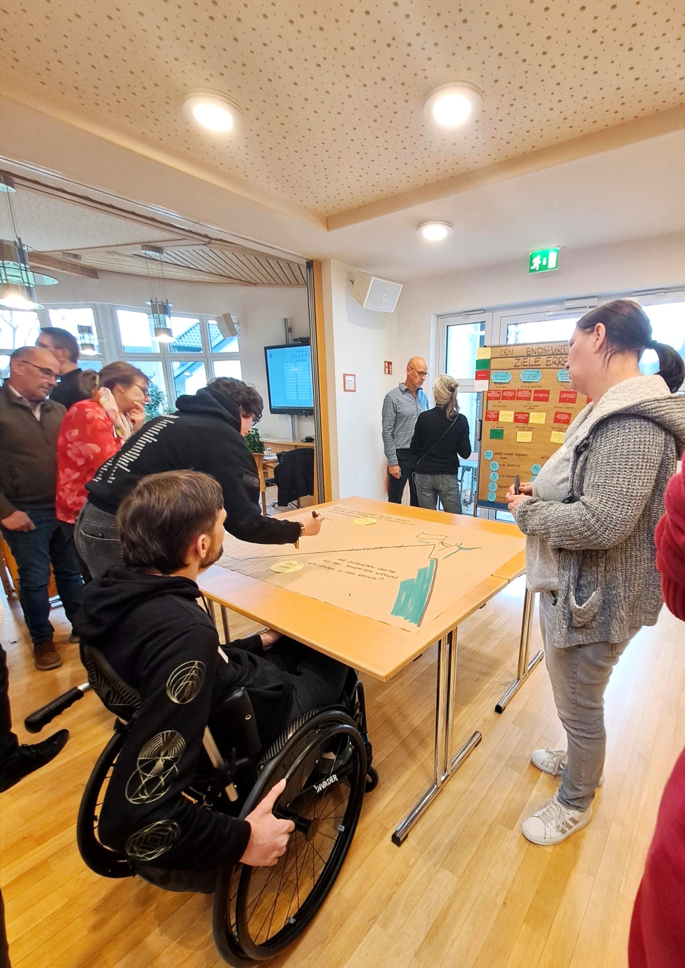 Eine Gruppe von Menschen versammelt sich um einen Tisch, um an einer Tafel zu schreiben und zu zeichnen. Einige stehen, während eine Person im Rollstuhl sitzt und an dem Projekt mitarbeitet.