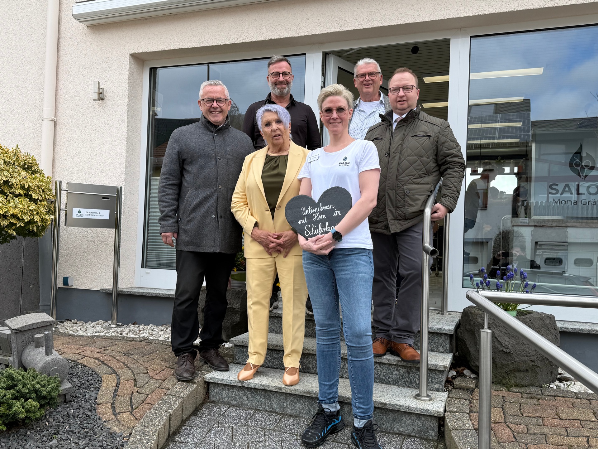 Die Aufnahme zeigt eine Gruppe von sechs Personen, die vor dem Friseursalon Mona Gräfen stehen. Die Personen sind auf verschiedenen Stufen einer Steintreppe positioniert. Von links nach rechts stehen fünf Männer und eine Frau in der Mitte. Die Männer tragen überwiegend dunkle, gedeckte Kleidung: Ein Mann links trägt einen grauen Mantel, der nächste einen schwarzen Hemdkragen, der dritte einen beige karierten Pullover, und der vierte Mann auf der rechten Seite trägt eine grüne Steppjacke. Die Frau in der Mitte trägt einen hellgelben Hosenanzug und hält in ihren Händen ein dunkles Herz aus Schiefer, auf dem in weißer Schrift "Unternehmen mit Herz im Schieferland" steht. Sie trägt ein weißes T-Shirt mit einem Logo. Ihr Haar ist kurz und blond, und sie trägt eine Sonnenbrille. Die gesamte Szene ist in neutralen Farben gehalten, mit dem hellen Gelb des Hosenanzugs als farblichem Akzent. Im Hintergrund sind Teile des Friseursalons sichtbar, einschließlich großer Fenster mit Reflexionen des Himmels und der Umgebung. Vor dem Gebäude sind ein Briefkasten und eine kleine Zierpflanze mit hellgrünen und gelben Blättern zu sehen. Links im Vordergrund befindet sich eine kleine graue Modell-Lokomotive. Die Treppe ist mit Steinen und Granit gepflastert. Die Komposition ist eine Frontalaufnahme der Gruppe, leicht von unten fotografiert, was die Personen prominent erscheinen lässt. Das Licht ist diffus, was auf einen bewölkten Tag hindeuten könnte.