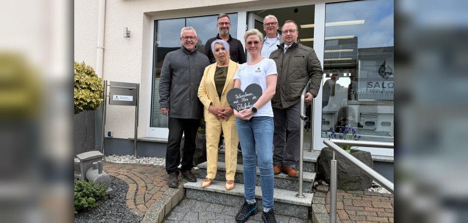 Die Aufnahme zeigt eine Gruppe von sechs Personen, die vor dem Friseursalon Mona Gräfen stehen. Die Personen sind auf verschiedenen Stufen einer Steintreppe positioniert. Von links nach rechts stehen fünf Männer und eine Frau in der Mitte. Die Männer tragen überwiegend dunkle, gedeckte Kleidung: Ein Mann links trägt einen grauen Mantel, der nächste einen schwarzen Hemdkragen, der dritte einen beige karierten Pullover, und der vierte Mann auf der rechten Seite trägt eine grüne Steppjacke. Die Frau in der Mitte trägt einen hellgelben Hosenanzug und hält in ihren Händen ein dunkles Herz aus Schiefer, auf dem in weißer Schrift "Unternehmen mit Herz im Schieferland" steht. Sie trägt ein weißes T-Shirt mit einem Logo. Ihr Haar ist kurz und blond, und sie trägt eine Sonnenbrille. Die gesamte Szene ist in neutralen Farben gehalten, mit dem hellen Gelb des Hosenanzugs als farblichem Akzent. Im Hintergrund sind Teile des Friseursalons sichtbar, einschließlich großer Fenster mit Reflexionen des Himmels und der Umgebung. Vor dem Gebäude sind ein Briefkasten und eine kleine Zierpflanze mit hellgrünen und gelben Blättern zu sehen. Links im Vordergrund befindet sich eine kleine graue Modell-Lokomotive. Die Treppe ist mit Steinen und Granit gepflastert. Die Komposition ist eine Frontalaufnahme der Gruppe, leicht von unten fotografiert, was die Personen prominent erscheinen lässt. Das Licht ist diffus, was auf einen bewölkten Tag hindeuten könnte.