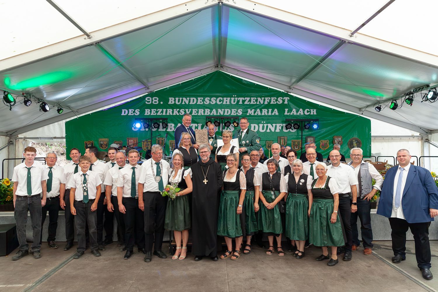Eine große Gruppe von Menschen, sowohl Männer als auch Frauen, steht unter einem Zeltdach für ein Gruppenfoto. Sie tragen Trachten und formelle Kleidung und hinter ihnen ist ein Banner mit der Aufschrift "98. BUNDESSCHÜTZENFEST DES BEZIRKSVERBANDES MARIA LAACH DÜNFUS". Abgebildet sind Personen von der St.-Sebastianus-Schützenbruderschaft Dünfus 1876 e. V. und Ehrengäste.