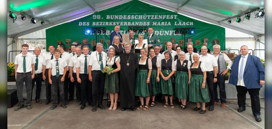 Gruppenfoto St.-Sebastianus-Schützenbruderschaft Dünfus 1876 e. V. mit Ehrengästen Eine große Gruppe von Menschen, sowohl Männer als auch Frauen, steht unter einem Zeltdach für ein Gruppenfoto. Sie tragen Trachten und formelle Kleidung und hinter ihnen ist ein Banner mit der Aufschrift "98. BUNDESSCHÜTZENFEST DES BEZIRKSVERBANDES MARIA LAACH DÜNFUS". Abgebildet sind Personen von der St.-Sebastianus-Schützenbruderschaft Dünfus 1876 e. V. und Ehrengäste.