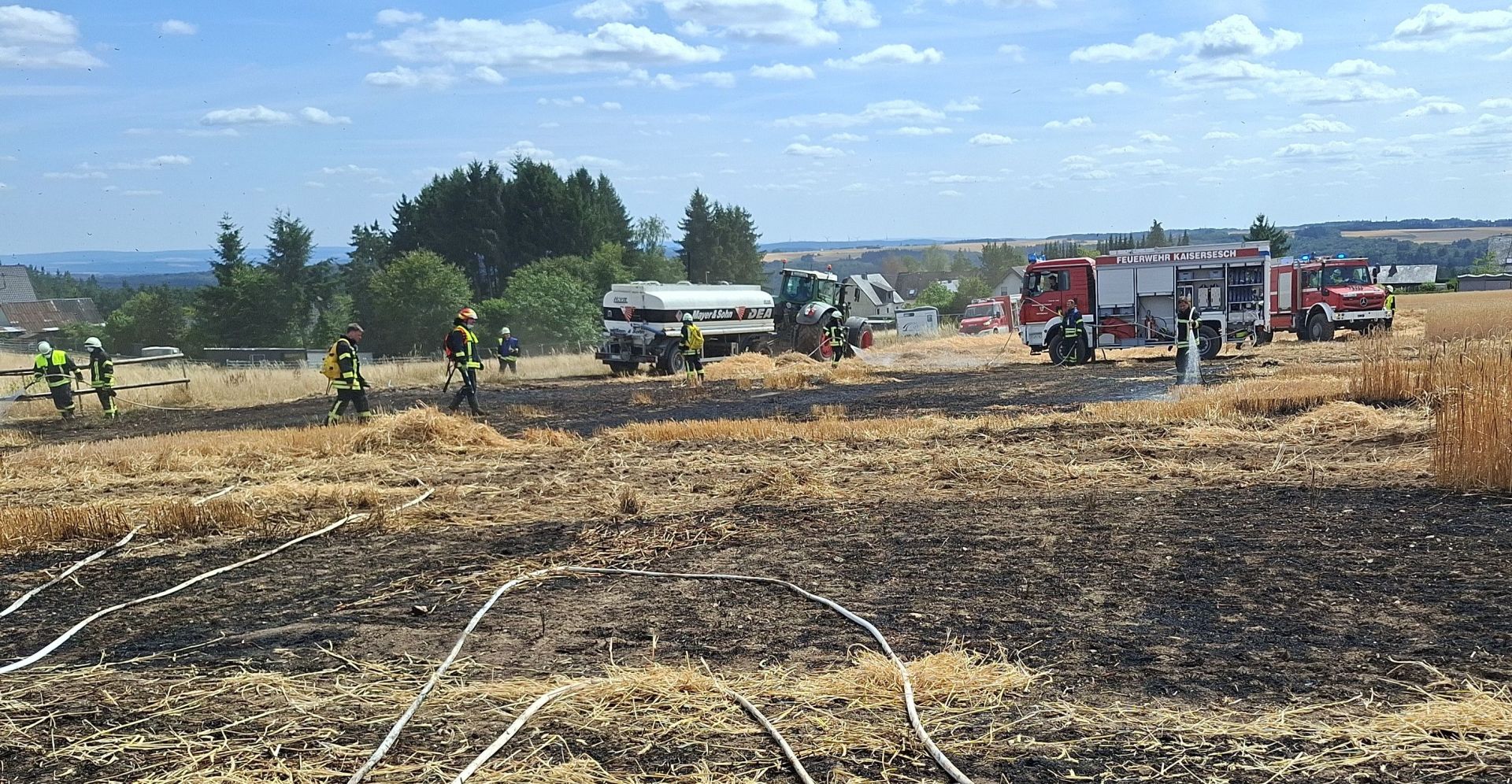 Die Szene zeigt ein Feld nach einem Brand, auf dem die Feuerwehr im Einsatz ist. Der Boden ist größtenteils schwarz verbrannt, mit vereinzelten Resten von Stroh und Getreideähren. Mehrere weiße Schläuche sind über das Feld verteilt, einige davon scheinen mit Wasser gefüllt zu sein. Im Hintergrund sind mehrere Einsatzfahrzeuge der Feuerwehr zu sehen, darunter rote Feuerwehrautos mit der Aufschrift „FEUERWEHR KAISERSESCH“. Auch ein Traktor und ein Tankwagen sind präsent. Mehrere Feuerwehrleute in gelben/schwarzen Einsatzanzügen und Schutzhelmen sind auf dem Feld tätig. Einige von ihnen scheinen mit den Schläuchen zu hantieren oder arbeiten an der Brandbekämpfung. Einer der Feuerwehrleute spritzt Wasser auf das noch brennende Feld. Das Wetter ist sonnig mit blauem Himmel und vereinzelten weißen Wolken. Am Horizont sind Hügel mit Bäumen und vereinzelten Gebäuden zu erkennen. 