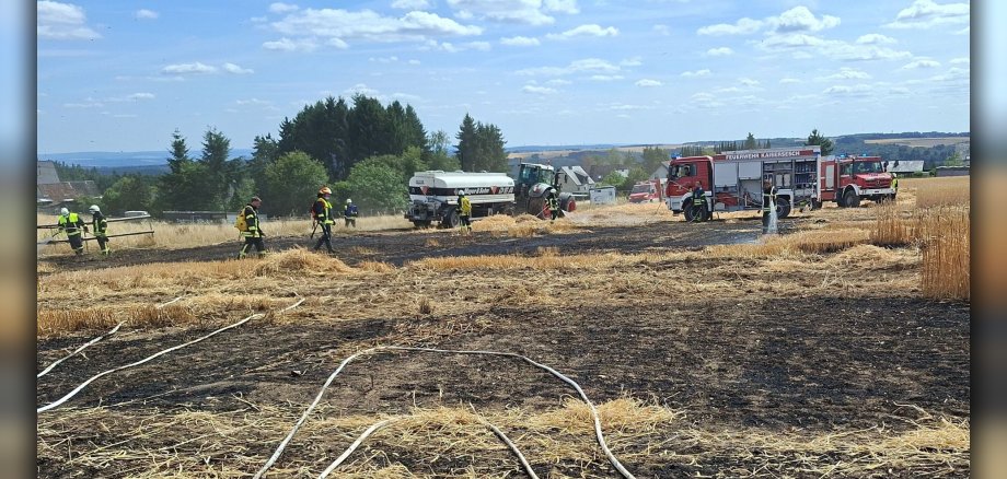 Einsatz der Feuerwehr bei einem Flächenbrand Die Szene zeigt ein Feld nach einem Brand, auf dem die Feuerwehr im Einsatz ist. Der Boden ist größtenteils schwarz verbrannt, mit vereinzelten Resten von Stroh und Getreideähren. Mehrere weiße Schläuche sind über das Feld verteilt, einige davon scheinen mit Wasser gefüllt zu sein. Im Hintergrund sind mehrere Einsatzfahrzeuge der Feuerwehr zu sehen, darunter rote Feuerwehrautos mit der Aufschrift „FEUERWEHR KAISERSESCH“. Auch ein Traktor und ein Tankwagen sind präsent. Mehrere Feuerwehrleute in gelben/schwarzen Einsatzanzügen und Schutzhelmen sind auf dem Feld tätig. Einige von ihnen scheinen mit den Schläuchen zu hantieren oder arbeiten an der Brandbekämpfung. Einer der Feuerwehrleute spritzt Wasser auf das noch brennende Feld. Das Wetter ist sonnig mit blauem Himmel und vereinzelten weißen Wolken. Am Horizont sind Hügel mit Bäumen und vereinzelten Gebäuden zu erkennen.