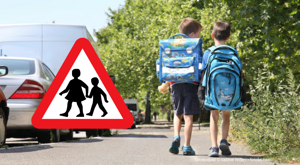 Das Bild zeigt zwei Kinder mit Schulranzen, die auf einem Bürgersteig entlanggehen. Im Vordergrund befindet sich ein dreieckiges Verkehrszeichen mit einem roten Rand und einem weißen Hintergrund, auf dem zwei schwarze Silhouetten von Kindern abgebildet sind, die Hand in Hand gehen. Es bedeutet „Achtung Kinder“ und warnt Autofahrer, dass hier vermehrt Kinder die Straße überqueren. Die beiden Kinder sind von hinten zu sehen, ihre Gesichter sind nicht sichtbar. Das linke Kind trägt einen blauen Schulranzen mit bunten Dinosaurier-Motiven, das rechte Kind einen ebenfalls blauen Schulranzen mit einem Schmetterlingsaufdruck. Im Hintergrund sind mehrere Autos am Straßenrand geparkt. Üppige grüne Bäume bilden eine dichte Kulisse am rechten und mittleren Bildrand. Das Bild vermittelt eine typische Alltagsszene im Kontext des Schulweges.  