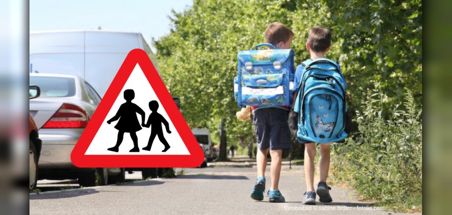 Das Bild zeigt zwei Kinder mit Schulranzen, die auf einem Bürgersteig entlanggehen. Im Vordergrund befindet sich ein dreieckiges Verkehrszeichen mit einem roten Rand und einem weißen Hintergrund, auf dem zwei schwarze Silhouetten von Kindern abgebildet sind, die Hand in Hand gehen. Es bedeutet „Achtung Kinder“ und warnt Autofahrer, dass hier vermehrt Kinder die Straße überqueren. Die beiden Kinder sind von hinten zu sehen, ihre Gesichter sind nicht sichtbar. Das linke Kind trägt einen blauen Schulranzen mit bunten Dinosaurier-Motiven, das rechte Kind einen ebenfalls blauen Schulranzen mit einem Schmetterlingsaufdruck. Im Hintergrund sind mehrere Autos am Straßenrand geparkt. Üppige grüne Bäume bilden eine dichte Kulisse am rechten und mittleren Bildrand. Das Bild vermittelt eine typische Alltagsszene im Kontext des Schulweges.  