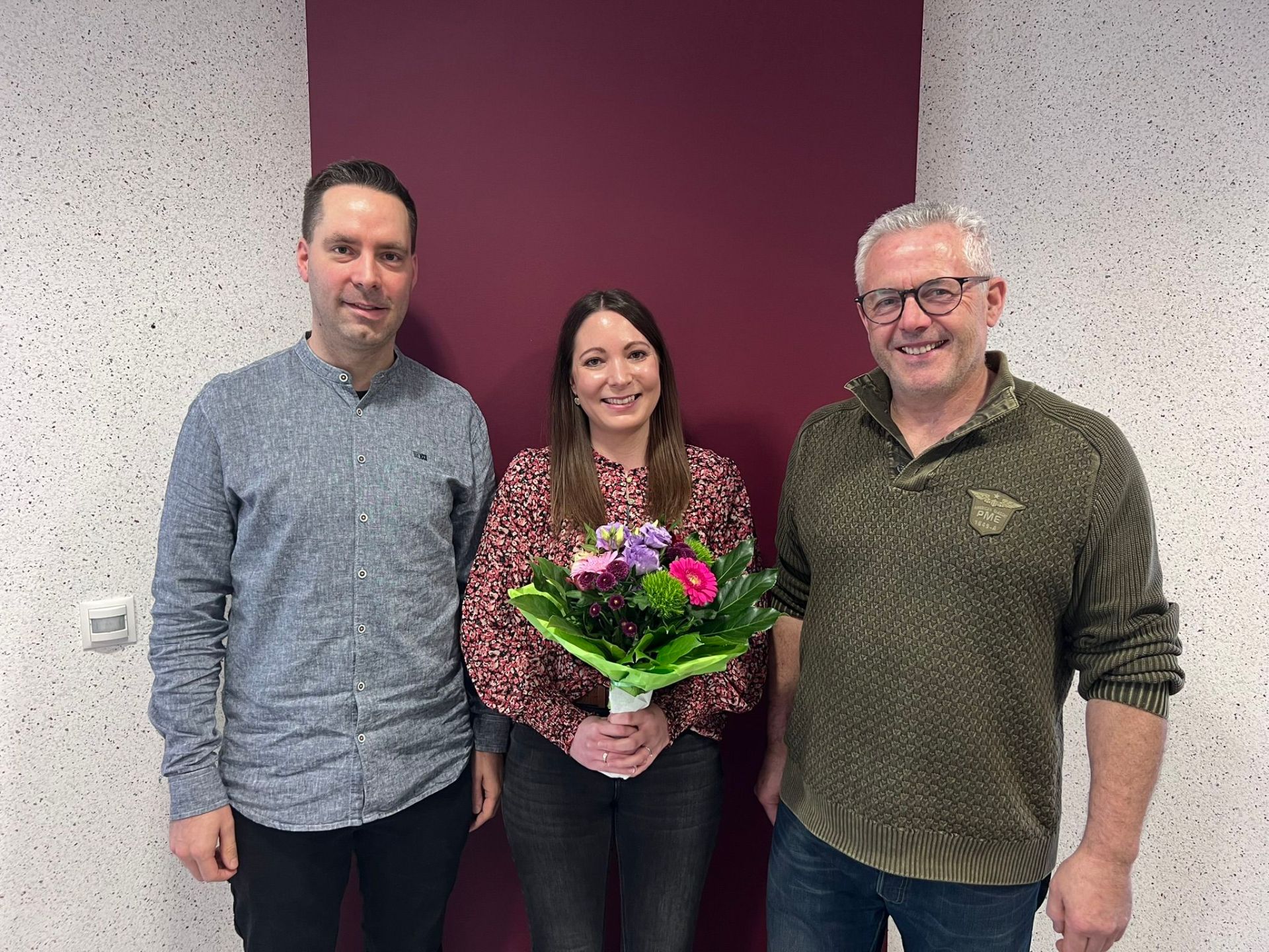 Das Bild zeigt drei Personen – zwei Männer und eine Frau – die nebeneinander stehen und in die Kamera lächeln. Die Frau in der Mitte hält einen bunten Blumenstrauß. Sie stehen vor einer Wand, die links und rechts von den Personen eine helle, gesprenkelte Textur aufweist, und im mittleren Bereich hinter den Personen dunkelrot ist.