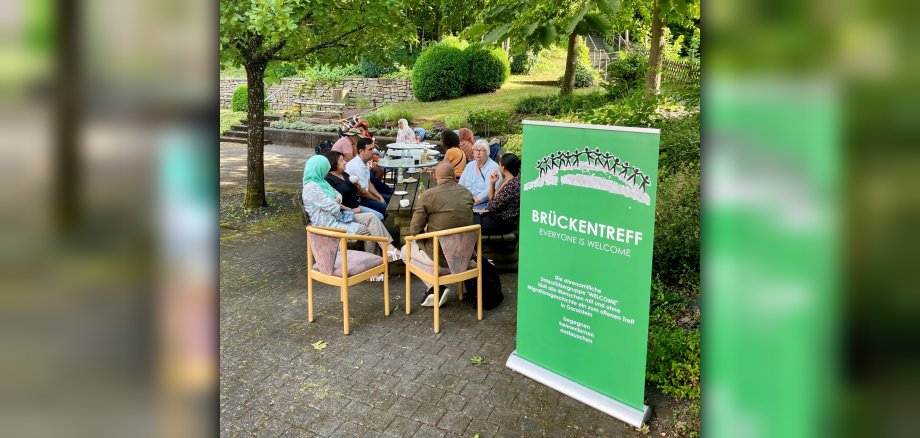Menschen sitzen in einem Park mit Bäumen und Sträuchern. Im Vordergrund steht ein Roll-up mit dem Hinweis auf den Brückentreff.