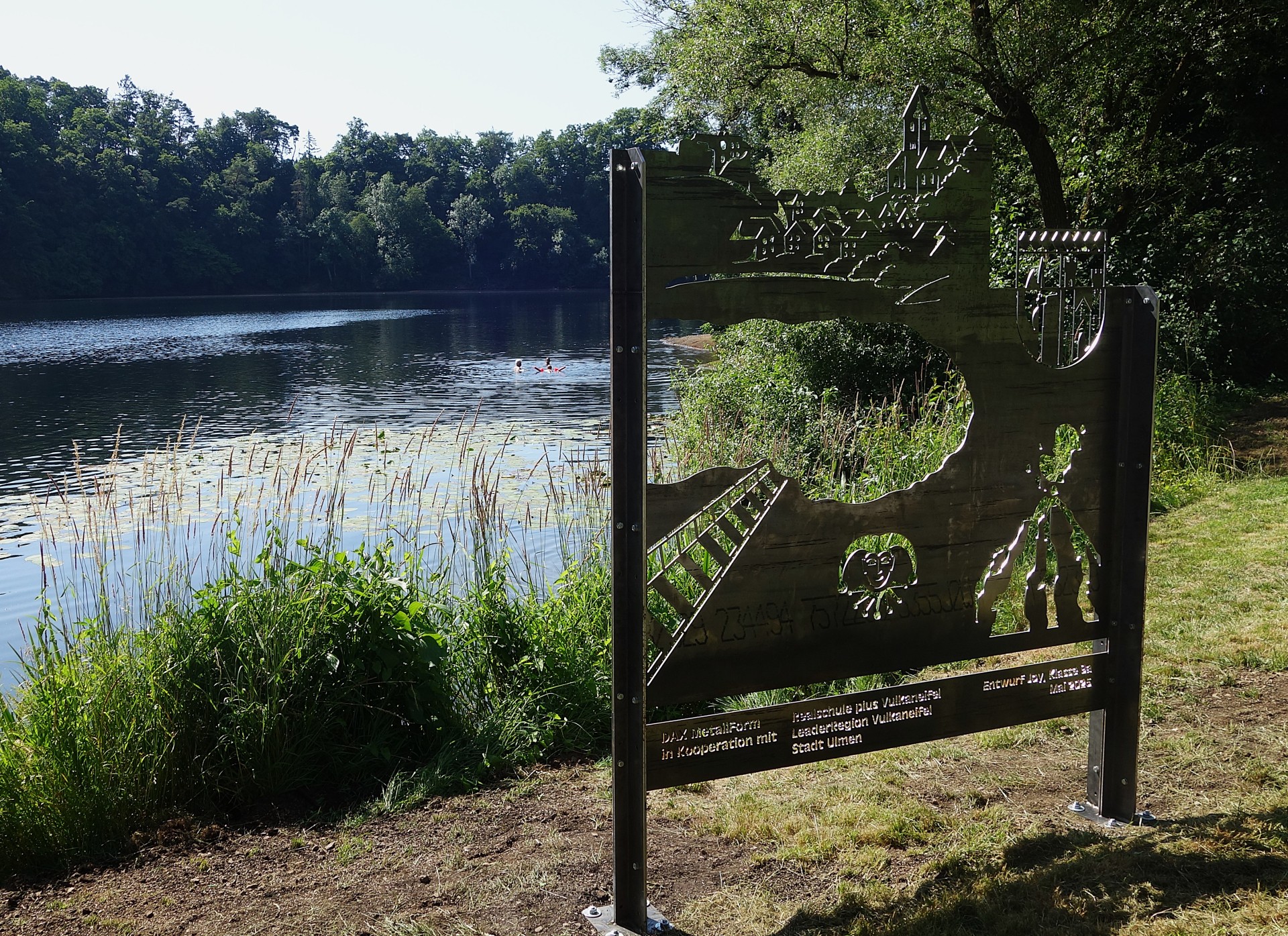 Fotopoint direkt am Ulmener Maar. Auf dem Foto sieht man das Ulmener Maar umrahmt von vielen grünen Bäumen. Im Vordergrund steht der Fotopoint, der Besucherinnen und Besuchern dazu einlädt, das Ulmener Maar als beeindruckende Kulisse für Erinnerungsfotos zu nutzen.