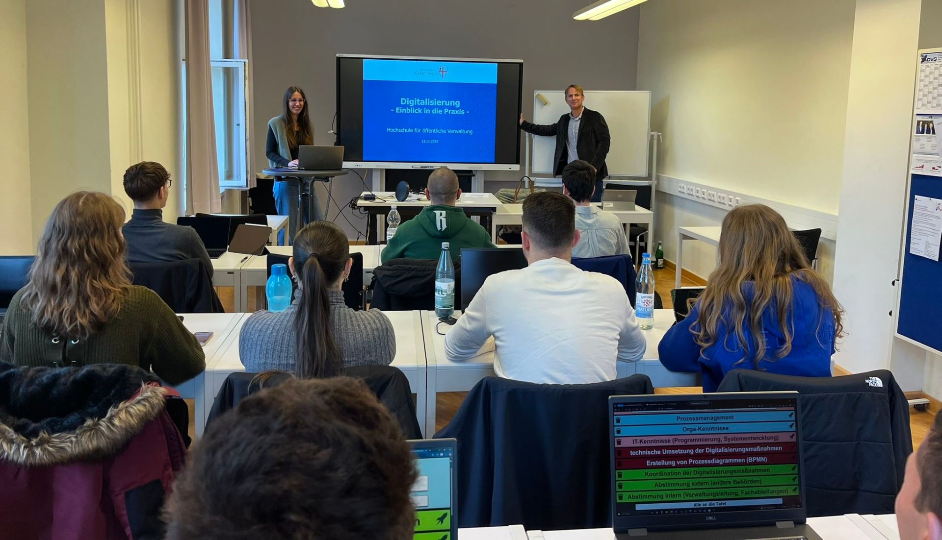 Unterricht an der Hochschule für öffentliche Verwaltung mit Studenten und Referenten. Es sind Frauen und Männer von hinten in einem Klassenraum zu sehen. Vorn stehen zwei Referenten, die eine Unterrichtseinheit zur Digitalisierung halten.