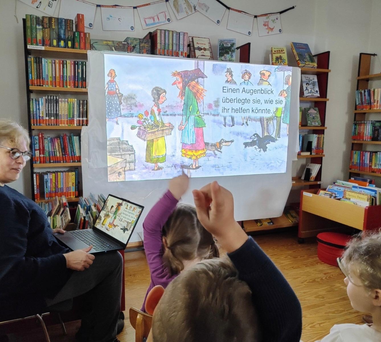 In einem Raum der katholischen öffentlichen Bücherei in Lutzerath sitzen Kinder und eine Dame. Die Dame präsentiert den Kindern über ein Bilderbuchkino, das auch auf einer Leinwand gezeigt wird, ein Bilderbuch. Die Kinder zeigen mit ihren Fingern auf, um etwas vorzulesen, was auf der Leinwand steht.