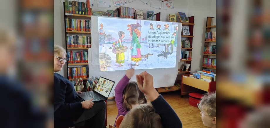 In einem Raum der katholischen öffentlichen Bücherei in Lutzerath sitzen Kinder und eine Dame. Die Dame präsentiert den Kindern über ein Bilderbuchkino, das auch auf einer Leinwand gezeigt wird, ein Bilderbuch. Die Kinder zeigen mit ihren Fingern auf, um etwas vorzulesen, was auf der Leinwand steht. In einem Raum der katholischen öffentlichen Bücherei in Lutzerath sitzen Kinder und eine Dame. Die Dame präsentiert den Kindern über ein Bilderbuchkino, das auch auf einer Leinwand gezeigt wird, ein Bilderbuch. Die Kinder zeigen mit ihren Fingern auf, um etwas vorzulesen, was auf der Leinwand steht.