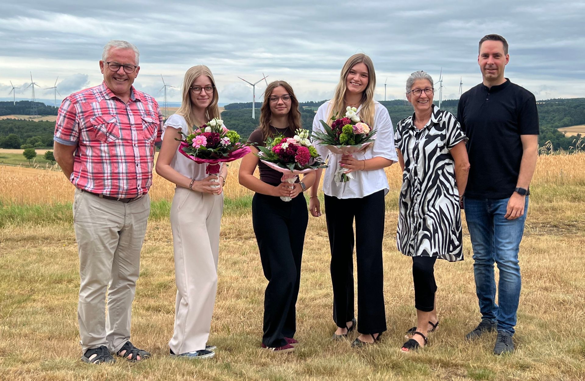 Gruppenfoto zur erfolgreich abgeschlossenen Ausbildung: Fünf Personen, drei Frauen und zwei Männer, stehen mit Blumensträußen in einem Feld vor Windrädern. Die Personen sind leger gekleidet und lächeln. Das Foto hat einen offiziellen Charakter und wirkt harmonisch.