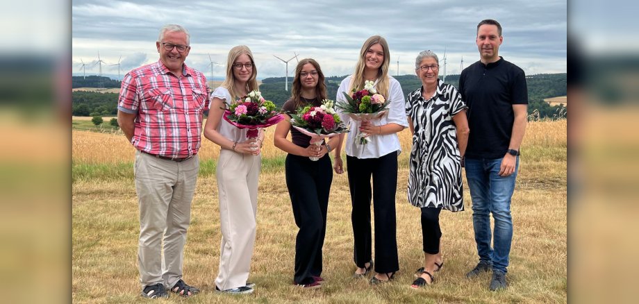 Gruppenfoto zur erfolgreich abgeschlossenen Ausbildung Gruppenfoto zur erfolgreich abgeschlossenen Ausbildung: Fünf Personen, drei Frauen und zwei Männer, stehen mit Blumensträußen in einem Feld vor Windrädern. Die Personen sind leger gekleidet und lächeln. Das Foto hat einen offiziellen Charakter und wirkt harmonisch.