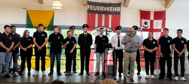 Gruppenfoto der Verpflichtungen Gruppenfoto der Verpflichtungen: 14 Männer und drei Frauen stehen bei der Verpflichtung auf einer Bühne. Die Männer und Frauen haben zum einen Teil Feuerwehranzüge an und zum anderen Teil legere Kleidung. Manche haben auch Poloshirts mit einem Feuerwehrabzeichen an. Im Vordergrund steht ein Mann, der ein Mikrofon in der Hand hält und von einem Zettel etwas abliest. Das Foto hat einen feierlichen Charakter.