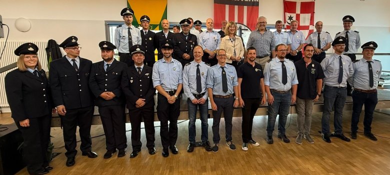 Gruppenfoto von den Ehrungen Gruppenfoto von den Ehrungen: 25 Männer und zwei Frauen stehen bei der Ehrung auf einer Bühne. Die Männer und Frauen haben zum einen Teil Feuerwehranzüge an und zum anderen Teil legere Kleidung. Manche haben auch Poloshirts mit einem Feuerwehrabzeichen an. Die Gruppe steht in zwei Reihen hintereinander. Das Foto hat einen feierlichen Charakter.