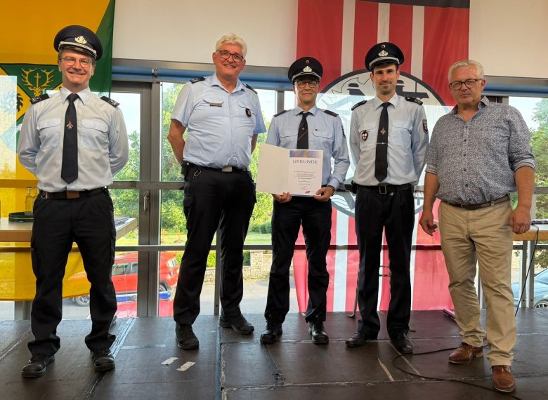 Gruppenfoto von den Ehrungen Gruppenfoto von den Ehrungen: fünf Männer stehen bei der Ehrung auf einer Bühne. Vier Männer haben eine blaue Hose und ein hellblaues Hemd an. Drei davon tragen eine Krawatte und eine Schirmmütze der Feuerwehr. Ein Mann hat eine beigefarbene Hose an und ein gemustertes Hemd. Das Foto hat einen feierlichen Charakter.