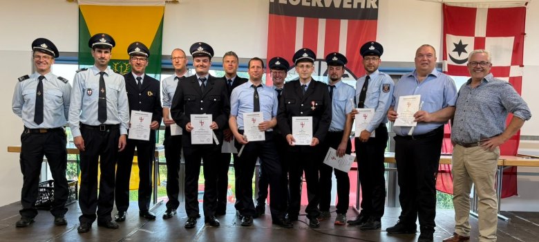 Gruppenfoto der Ernennungen der Wehrführer und ihrer Stellvertreter Gruppenfoto der Ernennungen der Wehrführer und ihrer Stellvertreter: Dreizehn Männer stehen bei der Ernennung auf der Bühne. Vier Männer haben Feuerwehranzüge an, acht Männer tragen blaue Hosen und ein hellblaues Hemd, davon haben sieben eine Krawatte an, ein Mann trägt eine beigefarbene Hose und ein gemustertes Hemd. Das Foto hat einen feierlichen Charakter.