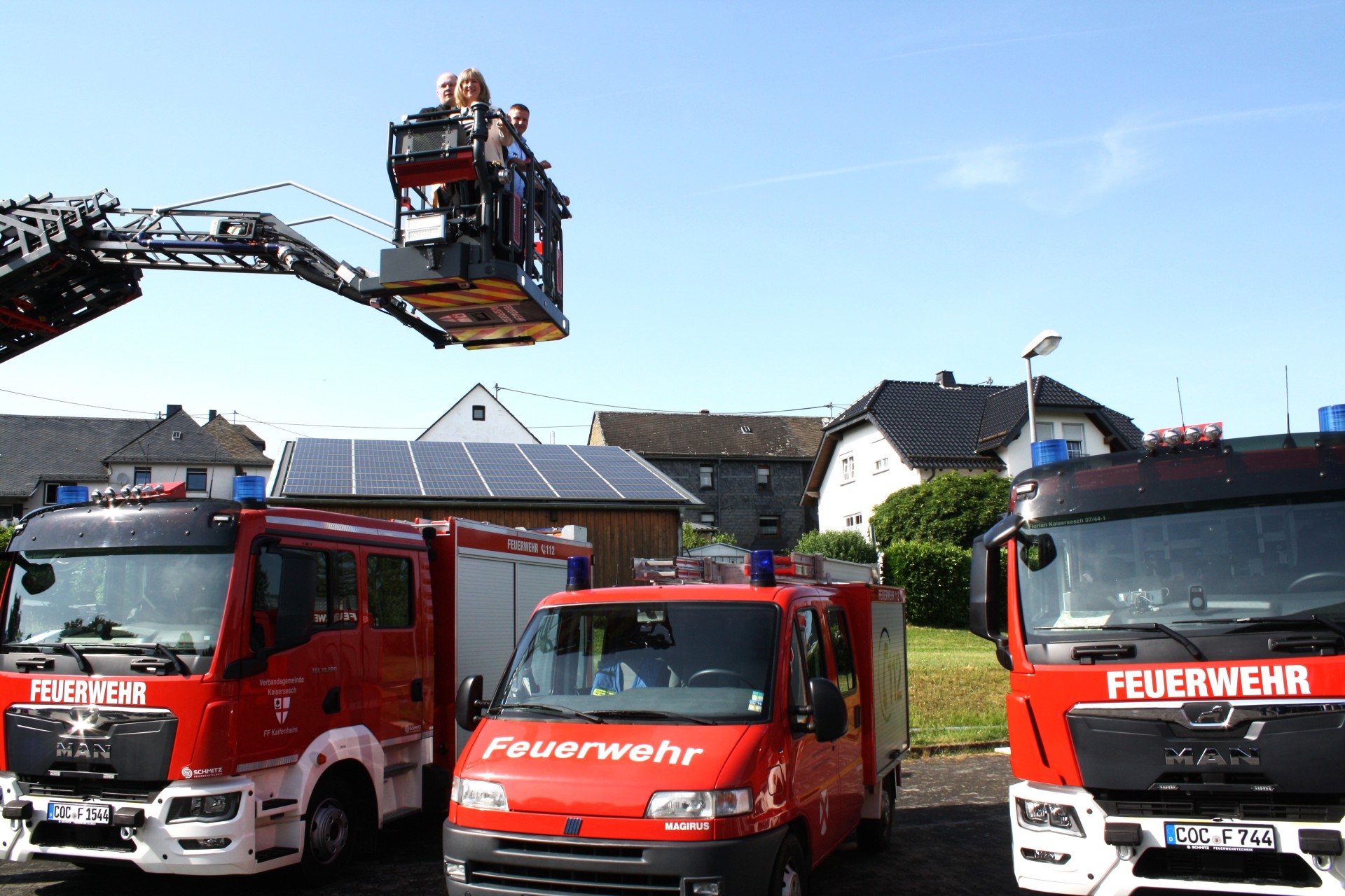 Einweihung der Feuerwehrfahrzeuge: Auf dem Foto unten links sind drei Feuerwehrfahrzeuge zu sehen. Darüber ist ein Kranausleger mit zwei Männern und einer Frau zu sehen. Im Hintergrund befinden sich eine Scheune mit Solardach und mehrere Häuser.