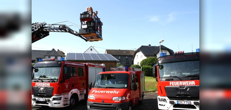 Einweihung der Feuerwehrfahrzeuge Einweihung der Feuerwehrfahrzeuge: Auf dem Foto unten links sind drei Feuerwehrfahrzeuge zu sehen. Darüber ist ein Kranausleger mit zwei Männern und einer Frau zu sehen. Im Hintergrund befinden sich eine Scheune mit Solardach und mehrere Häuser.