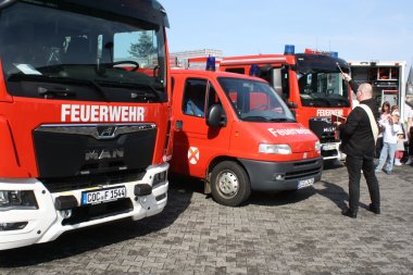 Einweihung der Feuerwehrfahrzeuge: Einweihung der Feuerwehrfahrzeuge: Drei Feuerwehrfahrzeuge stehen in einer Reihe, ganz rechts steht ein weiteres Feuerwehrfahrzeug, das nur von hinten und nur teilweise zu sehen ist. Am rechten Rand ist ein Mann im dunklen Anzug mit einer weißen Scherpe von hinten zu sehen. Weiter rechts sind noch zwei Frauen zu sehen.