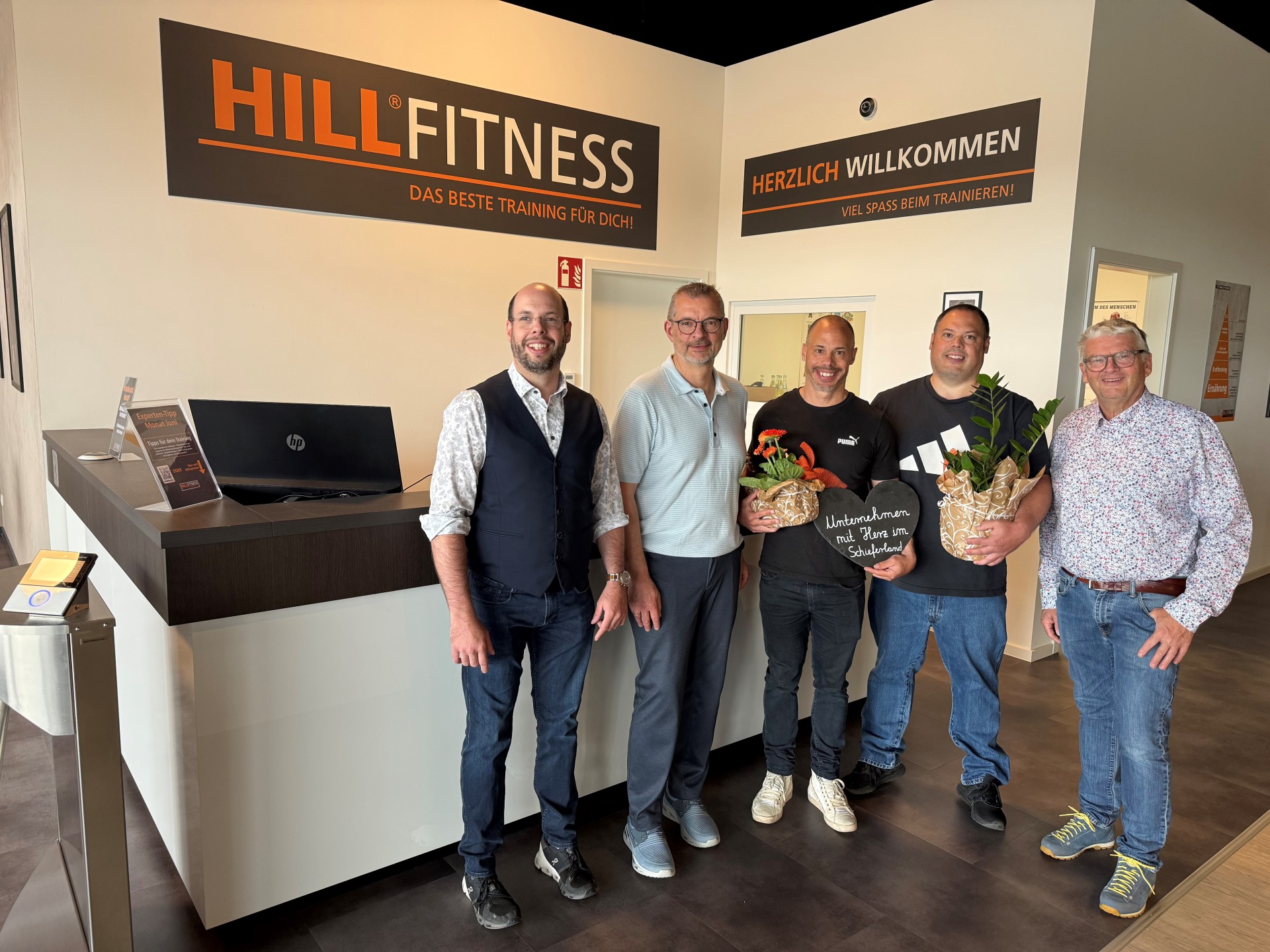 Gruppenfoto zur Eröffnung von Hill-Fitness in Kaisersesch: Auf dem Foto sind fünf Männer zu sehen, die leger gekleidet sind. Zwei Männer halten Blumen in den Händen. Im Hintergrund ist eine Theke zu sehen und über der Theke befinden sich zwei Schilder. Auf dem linken Schild steht „HillFitness - Das beste Training für dich“ und auf dem zweiten Schild steht „Herzlich Willkommen – Viel Spaß beim Trainieren“.