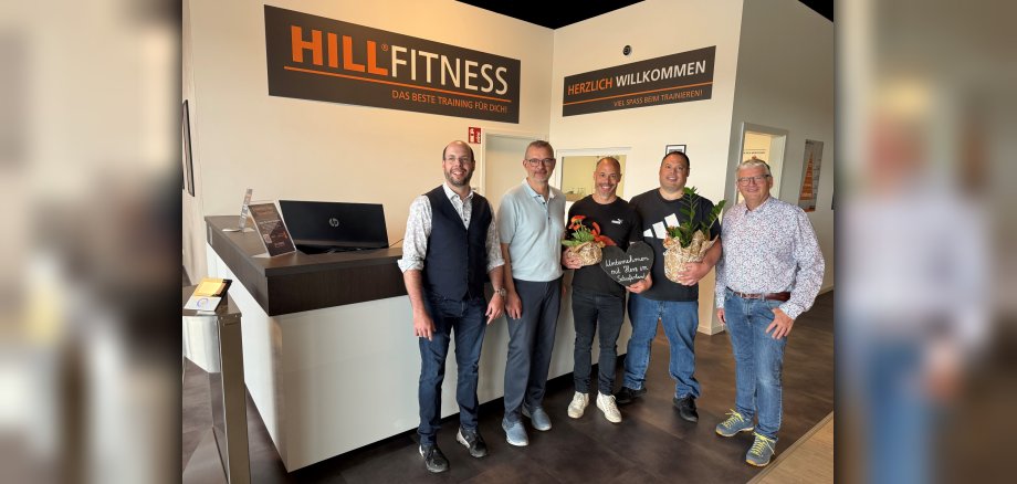 Gruppenfoto zur Eröffnung von Hill-Fitness in Kaisersesch Gruppenfoto zur Eröffnung von Hill-Fitness in Kaisersesch: Auf dem Foto sind fünf Männer zu sehen, die leger gekleidet sind. Zwei Männer halten Blumen in den Händen. Im Hintergrund ist eine Theke zu sehen und über der Theke befinden sich zwei Schilder. Auf dem linken Schild steht „HillFitness - Das beste Training für dich“ und auf dem zweiten Schild steht „Herzlich Willkommen – Viel Spaß beim Trainieren“.