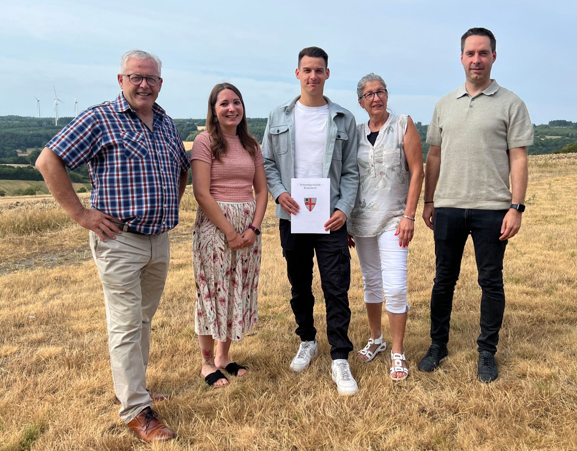 Die Aufnahme zeigt eine Gruppe von fünf Personen, die im Freien stehen, auf einem Feld mit trockenem, goldenem Gras. Im Hintergrund sind sanfte Hügel mit einigen Bäumen und im fernen Hintergrund Windräder zu sehen. Der Himmel ist bewölkt, aber hell. Von links nach rechts sind die Personen wie folgt: ein Mann mittleren Alters, der nach vorn blickt und lächelt. Er trägt eine karierte Kurzarmbluse in Blau und Rot über einer hellbraunen Hose und braunen Schnürschuhen. Seine Hände sind in die Hüften gestemmt. Er trägt eine Brille. Neben ihm steht eine Frau, die den Betrachter ebenfalls anlächelt. Sie trägt ein rosafarbenes Oberteil mit einer leichten Textur und einen langen, gemusterten Rock in Weiß mit roten und rosa Blütenmotiven. Sie trägt Sandalen und hat eine Tätowierung am rechten Bein. In der Mitte steht ein junger Mann, der ein Dokument hält. Er ist leicht nach links gedreht und blickt geradeaus. Er trägt ein graues Jackett über einem weißen T-Shirt und eine schwarze Cargohose. Seine Schuhe sind weiße Sneaker. Das Dokument, das er hält, ist weiß und hat ein Siegel mit einem roten und weißen Wappen sowie deutscher Schrift. Daneben steht eine ältere Frau, die leicht zur Seite blickt und lächelt. Sie trägt eine weiße, ärmellose Bluse mit einem floralen Muster und weiße 3/4-Hosen. Sie trägt eine Brille und weiße Sandalen. Ganz rechts steht ein Mann, der geradeaus blickt und leicht lächelt. Er trägt ein hellbraunes Poloshirt und eine schwarze Hose. Er trägt dunkle Schuhe und eine Armbanduhr. Die Komposition ist eine Gruppenaufnahme, bei der die Personen nebeneinander aufgereiht sind und den Großteil des unteren und mittleren Bildbereichs einnehmen. Der Hintergrund mit der Landschaft und den Windrädern ist leicht unscharf, was den Fokus auf die Personen lenkt. Die Farbpalette ist eher warm und natürlich, dominiert von den Gelb- und Brauntönen des Grases und den gedeckten Farben der Kleidung und der Landschaft. Das Wetter scheint mild und sommerlich zu sein.