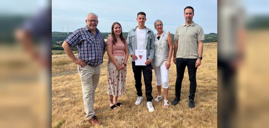 Gruppenfoto: Bürgermeister Albert Jung, Ausbildungsleiterin Carina Pauly, Lars Oster, Personalratsvorsitzende Petra Gotto und Büroleiter Mark Klasen Die Aufnahme zeigt eine Gruppe von fünf Personen, die im Freien stehen, auf einem Feld mit trockenem, goldenem Gras. Im Hintergrund sind sanfte Hügel mit einigen Bäumen und im fernen Hintergrund Windräder zu sehen. Der Himmel ist bewölkt, aber hell. Von links nach rechts sind die Personen wie folgt: ein Mann mittleren Alters, der nach vorn blickt und lächelt. Er trägt eine karierte Kurzarmbluse in Blau und Rot über einer hellbraunen Hose und braunen Schnürschuhen. Seine Hände sind in die Hüften gestemmt. Er trägt eine Brille. Neben ihm steht eine Frau, die den Betrachter ebenfalls anlächelt. Sie trägt ein rosafarbenes Oberteil mit einer leichten Textur und einen langen, gemusterten Rock in Weiß mit roten und rosa Blütenmotiven. Sie trägt Sandalen und hat eine Tätowierung am rechten Bein. In der Mitte steht ein junger Mann, der ein Dokument hält. Er ist leicht nach links gedreht und blickt geradeaus. Er trägt ein graues Jackett über einem weißen T-Shirt und eine schwarze Cargohose. Seine Schuhe sind weiße Sneaker. Das Dokument, das er hält, ist weiß und hat ein Siegel mit einem roten und weißen Wappen sowie deutscher Schrift. Daneben steht eine ältere Frau, die leicht zur Seite blickt und lächelt. Sie trägt eine weiße, ärmellose Bluse mit einem floralen Muster und weiße 3/4-Hosen. Sie trägt eine Brille und weiße Sandalen. Ganz rechts steht ein Mann, der geradeaus blickt und leicht lächelt. Er trägt ein hellbraunes Poloshirt und eine schwarze Hose. Er trägt dunkle Schuhe und eine Armbanduhr. Die Komposition ist eine Gruppenaufnahme, bei der die Personen nebeneinander aufgereiht sind und den Großteil des unteren und mittleren Bildbereichs einnehmen. Der Hintergrund mit der Landschaft und den Windrädern ist leicht unscharf, was den Fokus auf die Personen lenkt. Die Farbpalette ist eher warm und natürlich, dominiert von den Gelb- und Brauntönen des Grases und den gedeckten Farben der Kleidung und der Landschaft. Das Wetter scheint mild und sommerlich zu sein.