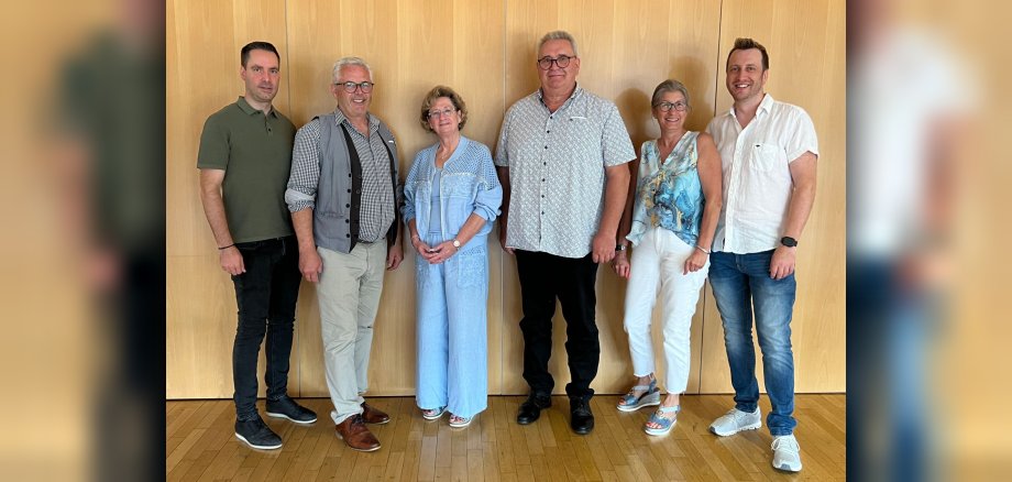 Sechs Personen stehen nebeneinander vor einer hellen Holzpaneelwand und lächeln in die Kamera. Sie tragen legere, sommerliche Kleidung.