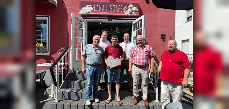 Gruppenfoto vor dem Lana Eiscafé in Kaisersesch Die Aufnahme zeigt sechs Männer vor einem roten Gebäude, das als "Lana Eiscafé" ausgeschildert ist. Sie stehen auf einer Steintreppe, die zur Eingangstür führt. Die Sonne scheint, was zu deutlichen Schattenwürfen führt. Im Hintergrund ist die Fassade des Eiscafés in einem kräftigen Rotton gehalten. Über dem Eingang hängt ein Schild mit dem Namen des Cafés in weißer Schrift, daneben Bilder von Kaffee und Eisbechern. Die Eingangstür und die beiden angrenzenden Fenster sind aus Glas und weiß. Links neben der Tür befindet sich ein Fenster mit Blumenkästen, die mit rosa Blumen und etwas Grünzeug geschmückt sind. Auf dem Sims davor steht ein kleiner weißer Tisch. Die sechs Männer sind in unterschiedlichen Altersgruppen und in unterschiedlicher Kleidung. Alle lächeln und scheinen posiert zu sein. Von links nach rechts: Der erste Mann von links hat graue Haare, trägt ein hellblaues Polohemd und Jeans. Seine Sonnenbrille hängt über dem Kragen. Der zweite Mann ist etwas jünger, trägt ein weißes Hemd und hat dunkle Haare. Er steht etwas im Schatten. Der dritte Mann, der jüngste in der Gruppe, trägt ein dunkelrotes Poloshirt und blaue Shorts. Er hält ein herzförmiges Schild mit deutscher Aufschrift "Unternehmern mit Herz im Sauerland" in den Händen. Er trägt Flip-Flops. Der vierte Mann hat graue Haare, trägt eine Brille, ein weißes Hemd mit einer leicht geöffneten Krawatte und eine dunkelbraune Hose. Der fünfte Mann von links ist deutlich älter, trägt ein kariertes Hemd in Rot- und Weißtönen, beige Hosen und braune Schuhe. Der sechste Mann, ganz rechts im Bild, hat kräftige Statur, kurze dunkle Haare und einen Bart. Er trägt ein knallrotes Poloshirt und helle Hosen. Seine Sonnenbrille trägt er auf dem Kopf. Die Beleuchtung ist hell und frontal, was die Farben des Gebäudes und der Kleidung hervorhebt. Die Komposition ist mittig, die Männer sind auf einer Linie arrangiert, was eine freundliche und einladende Atmosphäre schafft. Die grauen Steinstufen der Treppe bilden einen klaren Vordergrund und eine deutliche Trennung zum Hintergrund. Die Schatten auf den Stufen und den Männern geben dem Bild Tiefe.