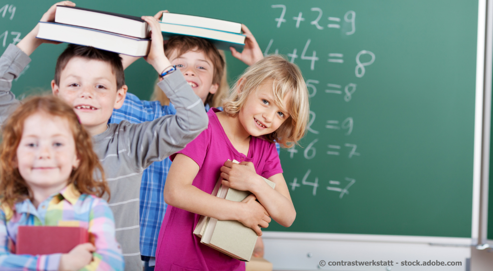 Kinder vor einer Schultafel, die Bücher auf dem Kopf und vor der Brust tragen: Im Vordergrund des Bildes sind vier Kinder vor einer grünen Schultafel zu sehen. Die Tafel ist mit mathematischen Gleichungen beschriftet, die in weißer Kreide geschrieben sind. Das vorderste Kind links ist ein Mädchen mit roten Haaren und Sommersprossen im Gesicht. Sie trägt ein kariertes Hemd in bunten Pastelltönen und hält ein rotes Buch in den Händen und lächelt leicht. Direkt hinter ihr steht ein Junge, der zwei Bücher auf seinem Kopf balanciert. Er hat braune Haare und trägt ein graues Langarmshirt mit horizontalen Streifen. Er grinst breit in die Kamera und wirkt sehr fröhlich. Neben dem Jungen, etwas weiter vorne und zur rechten Seite des Bildes, steht ein blondes Mädchen mit einem breiten Lächeln. Sie trägt ein pinkes T-Shirt und hält mehrere Bücher eng an ihre Brust gedrückt. Ein weiteres Kind, ist weiter oben zu sehen, man sieht nur ihren Oberkörper und ein Stück ihres blau-karierten Hemdes. Die Farben des Bildes sind lebendig; der Hintergrund mit der grünen Tafel und den mathematischen Gleichungen schafft eine schulische Atmosphäre. Die Komposition ist dynamisch durch die verschiedenen Tiefen der Kinder und ihre unterschiedlichen Posen und Gesichtsausdrücke. Der Fokus liegt auf den Kindern und ihren freudigen Ausdrücken, während die Tafel eher als dekoratives Element und zur Kontextualisierung dient. Das Bild strahlt eine positive und unbeschwerte Stimmung aus, die typisch für spielerische Momente im Schulalltag ist.  