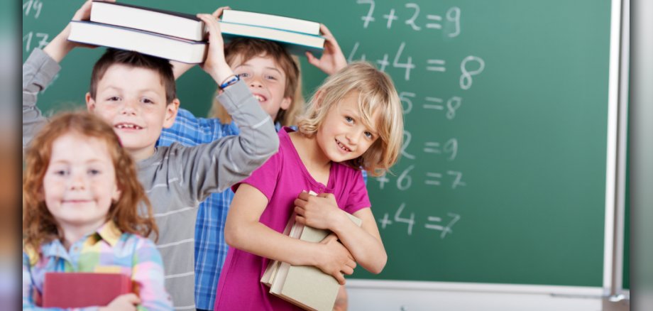 Kinder vor einer Schultafel, die Bücher auf dem Kopf und vor der Brust tragen. Kinder vor einer Schultafel, die Bücher auf dem Kopf und vor der Brust tragen: Im Vordergrund des Bildes sind vier Kinder vor einer grünen Schultafel zu sehen. Die Tafel ist mit mathematischen Gleichungen beschriftet, die in weißer Kreide geschrieben sind. Das vorderste Kind links ist ein Mädchen mit roten Haaren und Sommersprossen im Gesicht. Sie trägt ein kariertes Hemd in bunten Pastelltönen und hält ein rotes Buch in den Händen und lächelt leicht. Direkt hinter ihr steht ein Junge, der zwei Bücher auf seinem Kopf balanciert. Er hat braune Haare und trägt ein graues Langarmshirt mit horizontalen Streifen. Er grinst breit in die Kamera und wirkt sehr fröhlich. Neben dem Jungen, etwas weiter vorne und zur rechten Seite des Bildes, steht ein blondes Mädchen mit einem breiten Lächeln. Sie trägt ein pinkes T-Shirt und hält mehrere Bücher eng an ihre Brust gedrückt. Ein weiteres Kind, ist weiter oben zu sehen, man sieht nur ihren Oberkörper und ein Stück ihres blau-karierten Hemdes. Die Farben des Bildes sind lebendig; der Hintergrund mit der grünen Tafel und den mathematischen Gleichungen schafft eine schulische Atmosphäre. Die Komposition ist dynamisch durch die verschiedenen Tiefen der Kinder und ihre unterschiedlichen Posen und Gesichtsausdrücke. Der Fokus liegt auf den Kindern und ihren freudigen Ausdrücken, während die Tafel eher als dekoratives Element und zur Kontextualisierung dient. Das Bild strahlt eine positive und unbeschwerte Stimmung aus, die typisch für spielerische Momente im Schulalltag ist.