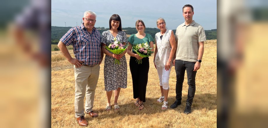 Das Bild zeigt fünf Personen, die in einer Reihe vor einem sonnigen, trockenen Feld stehen. Im Hintergrund sind sanfte Hügel mit einigen Bäumen und Windrädern zu sehen. Der Himmel ist leicht bewölkt und hellblau. Die Personen sind von links nach rechts angeordnet: Der Mann ganz links ist älter, trägt eine Brille, hat graue Haare und ein kariertes Hemd in Rot-, Blau- und Weißtönen. Seine Hose ist hellbeige und er trägt braune Lederschuhe. Er hat die Hände auf den Hüften. Die Frau neben ihm hat dunkle Haare mit Pony und trägt ein kurzärmeliges Kleid mit einem schwarzen Muster auf weißem Grund. Sie hält einen Blumenstrauß mit rosafarbenen, weißen und gelben Blumen in einer grünen Verpackung. Sie trägt helle Sandalen. Die Frau in der Mitte trägt ein smaragdgrünes Oberteil mit V-Ausschnitt und einen langen schwarzen Rock. Sie hält ebenfalls einen Blumenstrauß, ähnlich dem der Frau links, mit rosafarbenen, weißen und gelben Blumen. Sie trägt dunkle Sandalen. Die Frau neben ihr ist älter, trägt eine Brille und hat graue Haare. Sie trägt eine helle, gemusterte Tunika mit Blättermotiven und weiße 3/4-Hosen. Sie trägt weiße Sandalen. An ihrem linken Handgelenk ist ein Armband zu sehen. Der Mann ganz rechts trägt ein hellbeiges Poloshirt und eine dunkelgraue Jeans. Er hat dunkle Haare und trägt schwarze Schuhe. An seinem linken Handgelenk ist eine Smartwatch zu sehen. Der Vordergrund ist von trockenem, gelb-braunem Gras dominiert, das die Szene einrahmt und eine herbstliche oder sommerliche Atmosphäre vermittelt. Die Komposition ist so angelegt, dass alle fünf Personen sichtbar sind, wobei der Fokus auf ihnen liegt und der Hintergrund eine landschaftliche Ergänzung darstellt. Die Beleuchtung ist hell und gleichmäßig, was auf Tageslicht bei sonnigem Wetter hindeutet. Die Farben sind insgesamt warm, dominiert von Gelb- und Brauntönen im Gras, den grünen Blumensträußen und den hellen Kleidern der Personen.