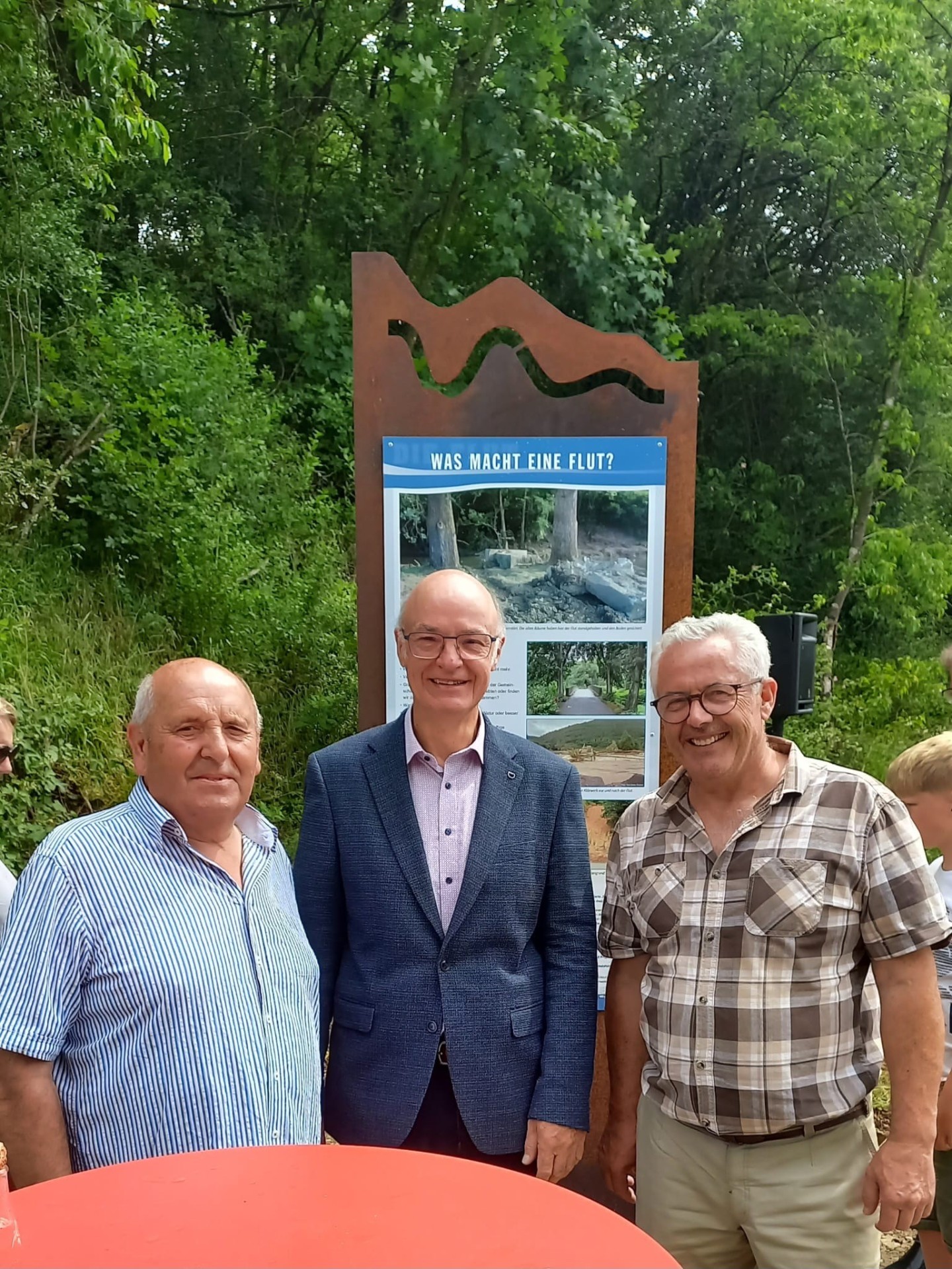 Drei ältere Männer (Ortsbürgermeister Ewald Neisz von Insul (links), Robert Reuter von Dümpelfeld (Mitte), Bürgermeister Albert Jung (rechts)) stehen in der Nähe eines Informationsschildes in einer baumbestandenen Umgebung. Im Vordergrund ist die Oberseite eines runden, roten Tisches zu sehen. Die drei Männer stehen nebeneinander im mittleren Bereich des Bildes. Der Mann ganz links trägt ein blau-weiß gestreiftes Hemd und lächelt in die Kamera. Er hat eine Glatze und helle Haut. Der Mann in der Mitte trägt einen dunkelblauen Anzug über einem hellrosa Hemd mit einem Muster und hat eine Brille auf. Er lächelt ebenfalls. Seine Haut ist blass. Der Mann ganz rechts trägt ein kurzärmeliges, kariertes Hemd in Braun- und Beigetönen über einer hellbraunen Hose. Auch er trägt eine Brille und lächelt. Seine Haut ist ebenfalls hell. Hinter den Männern steht ein großes, ornamentales Informationsschild. Das Schild hat eine rostige Metallkonstruktion an der Oberseite, die in einem wellenartigen Muster geformt ist. Darunter befindet sich eine blaue Tafel mit weißer Schrift. Über einem Bild, das einen Baumstumpf und Steine zeigt, steht die Frage "WAS MACHT EINE FLUT?". Darunter sind mehrere kleinere Bilder und Text zu sehen, die die Auswirkungen von Überschwemmungen darstellen. Der Hintergrund des Bildes ist üppig grün, dominiert von dichten Bäumen und Büschen, was auf eine sommerliche oder frühlingshafte Szenerie hindeutet. Das Licht ist hell, aber diffus, was darauf schließen lässt, dass es vielleicht bewölkt ist oder die Sonne von den Blättern verdeckt wird. Die Komposition ist auf die drei Männer und das Informationsschild fokussiert, mit dem natürlichen Grün als Hintergrund. Im rechten Teil des Bildes sind noch weitere Personen, darunter ein Kind, schemenhaft zu erkennen, ebenso wie eine schwarze Säule, die möglicherweise eine Lautsprecherbox ist.