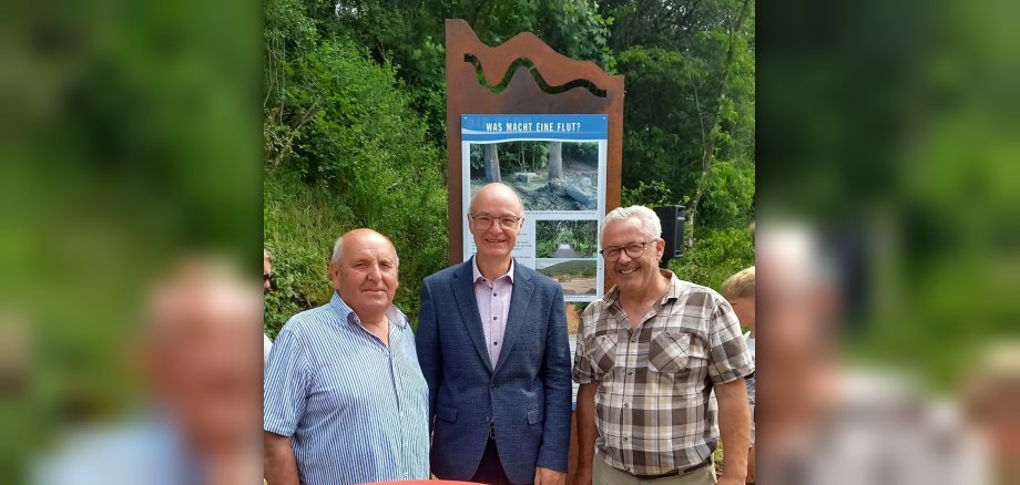 Gruppenfoto mit drei Männern Drei ältere Männer (Ortsbürgermeister Ewald Neisz von Insul (links), Robert Reuter von Dümpelfeld (Mitte), Bürgermeister Albert Jung (rechts)) stehen in der Nähe eines Informationsschildes in einer baumbestandenen Umgebung. Im Vordergrund ist die Oberseite eines runden, roten Tisches zu sehen. Die drei Männer stehen nebeneinander im mittleren Bereich des Bildes. Der Mann ganz links trägt ein blau-weiß gestreiftes Hemd und lächelt in die Kamera. Er hat eine Glatze und helle Haut. Der Mann in der Mitte trägt einen dunkelblauen Anzug über einem hellrosa Hemd mit einem Muster und hat eine Brille auf. Er lächelt ebenfalls. Seine Haut ist blass. Der Mann ganz rechts trägt ein kurzärmeliges, kariertes Hemd in Braun- und Beigetönen über einer hellbraunen Hose. Auch er trägt eine Brille und lächelt. Seine Haut ist ebenfalls hell. Hinter den Männern steht ein großes, ornamentales Informationsschild. Das Schild hat eine rostige Metallkonstruktion an der Oberseite, die in einem wellenartigen Muster geformt ist. Darunter befindet sich eine blaue Tafel mit weißer Schrift. Über einem Bild, das einen Baumstumpf und Steine zeigt, steht die Frage "WAS MACHT EINE FLUT?". Darunter sind mehrere kleinere Bilder und Text zu sehen, die die Auswirkungen von Überschwemmungen darstellen. Der Hintergrund des Bildes ist üppig grün, dominiert von dichten Bäumen und Büschen, was auf eine sommerliche oder frühlingshafte Szenerie hindeutet. Das Licht ist hell, aber diffus, was darauf schließen lässt, dass es vielleicht bewölkt ist oder die Sonne von den Blättern verdeckt wird. Die Komposition ist auf die drei Männer und das Informationsschild fokussiert, mit dem natürlichen Grün als Hintergrund. Im rechten Teil des Bildes sind noch weitere Personen, darunter ein Kind, schemenhaft zu erkennen, ebenso wie eine schwarze Säule, die möglicherweise eine Lautsprecherbox ist.