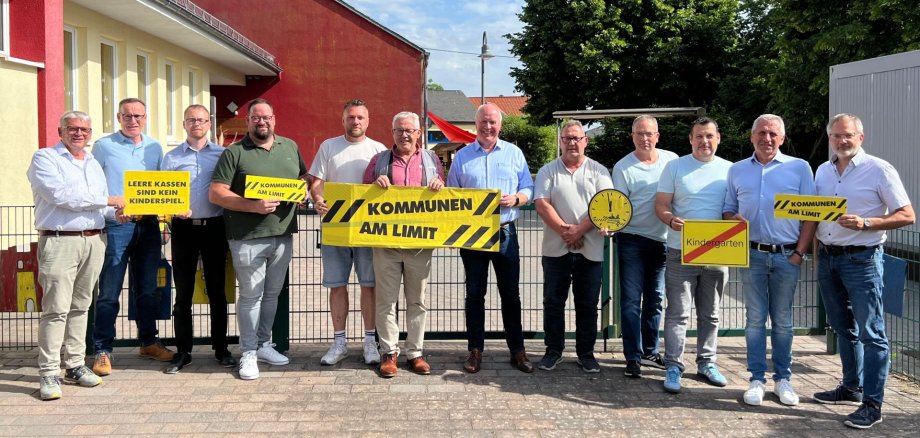 Eine Gruppe von elf Männern steht nebeneinander vor einer roten Gebäudefassade und einem Zaun. Sie halten verschiedene Schilder hoch, die auf ihre Anliegen aufmerksam machen sollen. Die Schilder sind gelb mit schwarzer Schrift und haben teilweise schwarze Streifen an den Rändern, die ein Gefahrensymbol andeuten. Von links nach rechts sind die Schilder wie folgt zu erkennen: 1. Ein Mann hält ein gelbes Schild mit der Aufschrift „LEERE KASSEN SIND KEIN KINDERSPIEL“. 2. Ein Mann hält ein gelbes Schild mit schwarzen Streifen und der Aufschrift „KOMMUNEN AM LIMIT“. 3. Ein weiterer Mann hält dasselbe „KOMMUNEN AM LIMIT“ Schild. 4. Vor dem fünften Mann von links, der ebenfalls ein „KOMMUNEN AM LIMIT“ Schild hält, sieht man ein gelbes Schild mit der Aufschrift „Kindergarten“. Dieses Schild hat einen roten Streifen am oberen und unteren Rand, ähnlich einem Ortsschild. 5. Neben dem „Kindergarten“ Schild steht ein Mann, der ein großes gelbes Schild mit schwarzen Streifen hochhält, auf dem „KOMMUNEN AM LIMIT“ steht. 6. Der vorletzte Mann von links hält ein gelbes Schild mit schwarzen Streifen und der Aufschrift „KOMMUNEN AM LIMIT“. 7. Der allerletzte Mann von links hält ebenfalls ein „KOMMUNEN AM LIMIT“ Schild. Zusätzlich zu den Schildern hält der Mann, der das „Kindergarten“ Schild in der Hand hält, eine gelbe Scheibe mit einer schwarzen Zeigeruhr, die ungefähr 2 Minuten vor 12:00 Uhr steht. Auf der Uhr sind stilisierte Gebäude zu sehen. Die Männer tragen überwiegend Hemden und Jeans, einige auch Poloshirts. Die Kleidung ist größtenteils in Blau-, Grau- und Weißtönen gehalten. Die Stimmung wirkt ernst, aber auch entschlossen. Die Männer sind verschieden alt, von mittleren Jahren bis ins höhere Alter. Die Umgebung im Hintergrund besteht aus einer rot-gelben Fassade eines Gebäudes, es ist das Kindergartengebäude St. Nikolaus in Kaifenheim. Dahinter sind Bäume und ein blauer Himmel mit einigen Wolken zu sehen. Ein Metallzaun trennt die Gruppe von dem Gebäude. Die Aufnahme ist im Freien bei Tageslicht entstanden. Die Komposition ist eine Gruppenaufnahme von vorn, wobei die Männer dicht nebeneinander aufgereiht sind und ihre Schilder sichtbar sind.