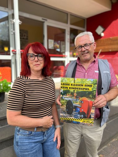 Ein Mann und eine Frau stehen nebeneinander vor einem Gebäude. Die Frau, links im Bild, hat feuerrote Haare, die ihr bis zu den Schultern reichen und eine Ponyfrisur haben. Sie trägt eine Brille mit schwarzem Rahmen und eine gestreifte Strickjacke mit horizontalen Streifen in Beige und Schwarz. Ihre Hose ist aus verwaschenem Denim und sie trägt einen braunen Gürtel mit einer runden Schnalle. Die Frau blickt direkt in die Kamera und ihre Mimik ist ernst. Der Mann, rechts im Bild, hat kurze, graue Haare und trägt eine Brille mit dunklem, rundem Rahmen. Er hat eine graue Weste über einem rosafarbenen Hemd mit kurzen Ärmeln und offenes Kragen. Er hält ein Plakat hoch, auf dem ein Spielgerät zu sehen ist. Auf dem Plakat steht in großen gelben Buchstaben "LEERE KASSEN SIND KEIN KINDERSPIEL.". Darunter steht in kleineren Buchstaben "KOMMUNEN AM LIMIT". Der Mann lächelt in die Kamera. Der Hintergrund zeigt den Eingang des Kindergartens St. Nikolaus in Kaifenheim. Auf der linken Seite sind ein paar grüne Pflanzen zu sehen, die in einem Topf stehen. Das Licht im Bild ist hell und die Farben sind lebendig. Die Komposition ist frontale und die beiden Personen nehmen einen großen Teil des Bildes ein. Der Fokus liegt auf den Personen und dem Plakat, das sie hochhalten.