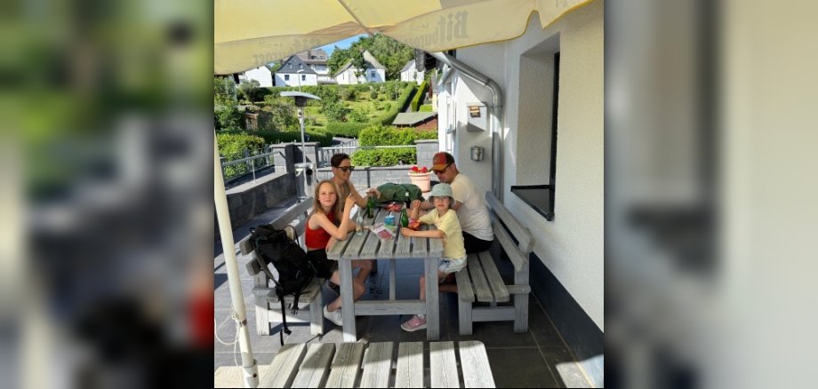 Das Bild zeigt eine Familie, die an einem Holztisch im Freien sitzt. Sie befinden sich auf einer Terrasse des Brohlbachstübchens in Gamlen unter einem großen, gelben Sonnenschirm. Der Hintergrund zeigt eine hügelige Landschaft mit mehreren kleinen Häusern und üppiger grüner Vegetation, die auf einen sonnigen Tag hindeutet. Im Vordergrund des Bildes stehen eine Familie von vier Personen, zwei Erwachsene und zwei Kinder, um einen grauen Holztisch. Sie scheinen sich in einer entspannten und geselligen Atmosphäre zu befinden. Der Tisch ist mit einigen Flaschen, möglicherweise Getränken, und vielleicht ein paar Snacks belegt. Links auf dem Bild sitzt ein Mädchen mit langen, hellen Haaren, das ein rotes Top trägt. Neben ihr auf der Bank liegt ein schwarzer Rucksack. Sie schaut leicht nach links, aber ihre Aufmerksamkeit scheint dem Tisch zu gelten. Auf der anderen Seite des Tisches, rechts vom Mädchen, sitzt ein jüngeres Kind mit einem hellblauen oder grauen Hut und einem gelben T-Shirt. Dieses Kind schaut direkt in die Kamera. Neben ihm sitzt ein Erwachsener, ein Mann, der eine Kappe und ein weißes T-Shirt trägt. Er schaut nach rechts. Eine Frau, die wahrscheinlich die Mutter ist, sitzt ebenfalls am Tisch und schaut nach vorn. Sie trägt eine Sonnenbrille und ein helles Oberteil. Die Farben im Bild sind überwiegend warm und hell, was die sonnige Wetterstimmung unterstreicht. Das Gelb des Sonnenschirms dominiert den oberen Teil des Bildes und schafft einen starken Kontrast zum blauen Himmel und dem grünen Hintergrund. Das Grau der Holztische und Bänke wirkt natürlich und fügt sich gut in die Umgebung ein. Die Kleidung der Personen ist ebenfalls hell und sommerlich. Die Komposition des Bildes ist etwas von oben aufgenommen und zeigt die Gruppe von der Seite. Der Sonnenschirm nimmt einen erheblichen Teil des oberen Bildbereichs ein und erzeugt Schatten, die für eine angenehme Atmosphäre sorgen. Die Landschaft im Hintergrund ist in leicht unscharfen Details gehalten, was den Fokus auf die Familie lenkt. Es gibt eine gewisse Tiefe im Bild, die durch die Staffelung der Familie, des Tisches und der dahinterliegenden Landschaft entsteht. Man sieht auch weitere Holztische und Bänke im Vordergrund des Brohlbachstübchens. Rechts im Bild ist die Wand des Brohlbachstübchens zu sehen, mit einem Metallrohr und einem Lichtschalter an der Fassade. Insgesamt vermittelt das Bild eine friedliche und angenehme Szene eines Familienausflugs an einem schönen Sommertag.
